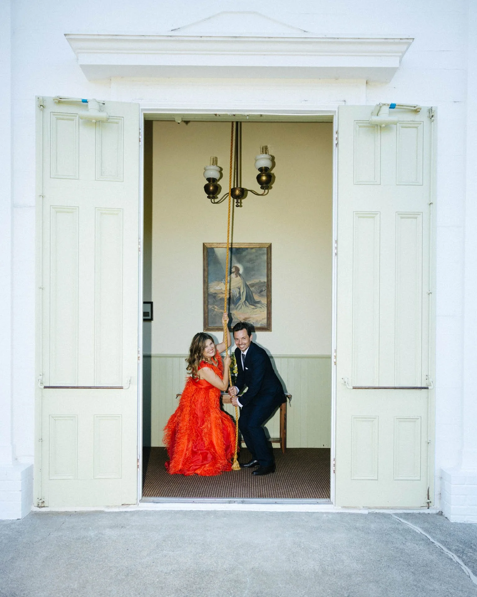 A couple dressed in formal attire, the woman in a red dress and the man in a black tuxedo, are inside a room with open white doors, holding a rope together and smiling.