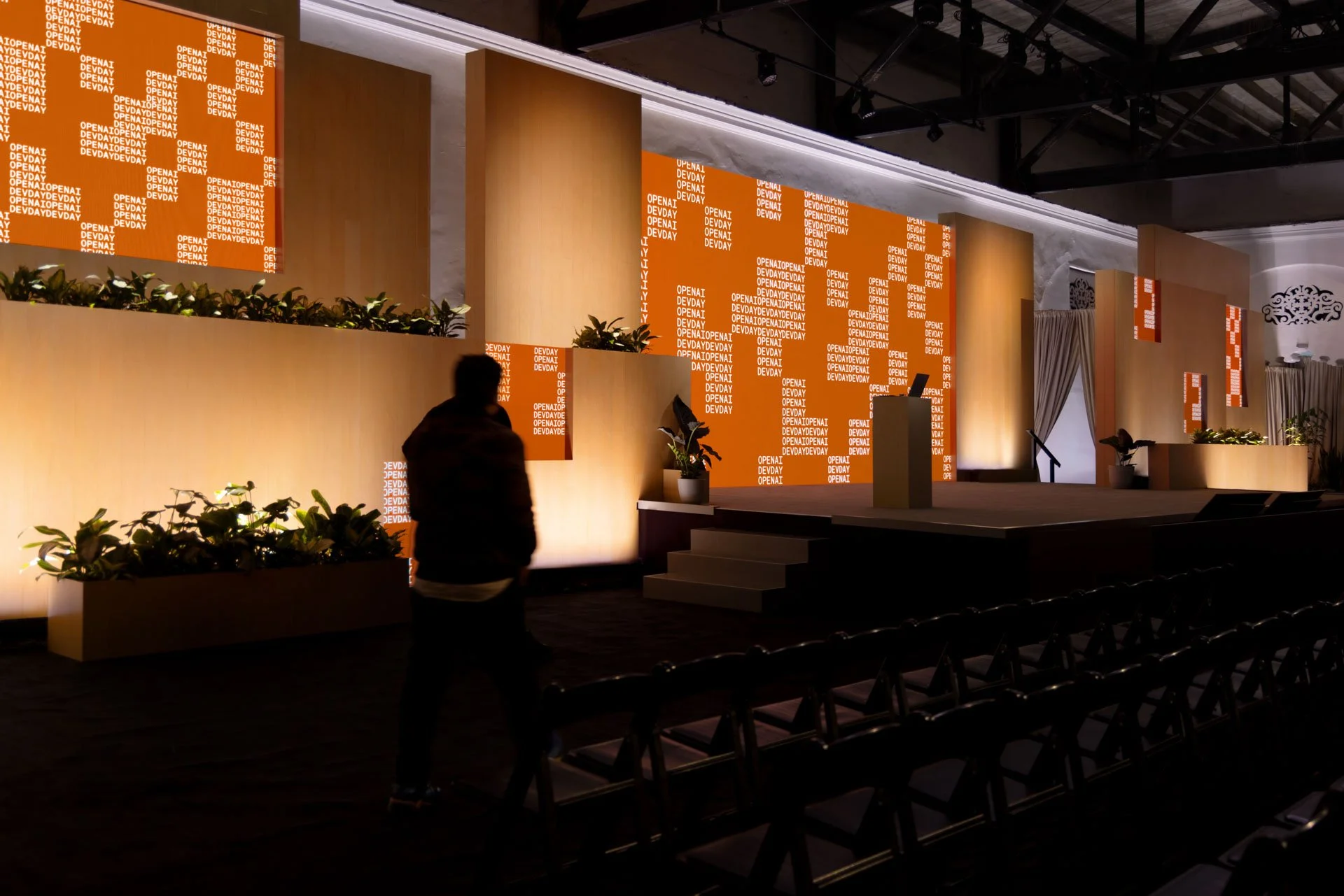An empty stage with a podium, illuminated by warm lighting, with large orange screens displaying white text that reads 'OPENAI.' There are potted plants and curtains on the sides, and a person is walking in front of the stage in silhouette.