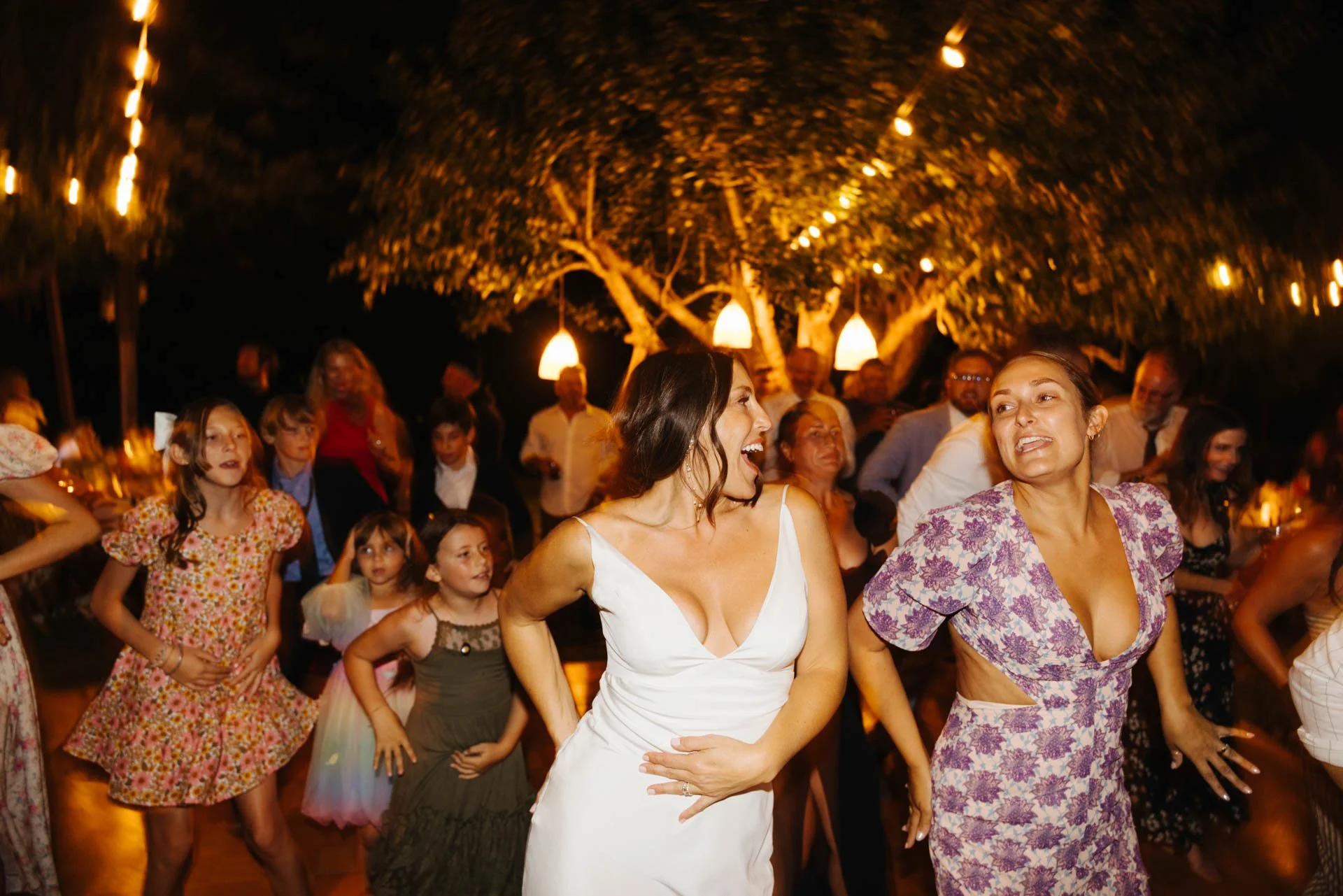 People dancing at a wedding reception outdoors during the night, with string lights and a large tree in the background.