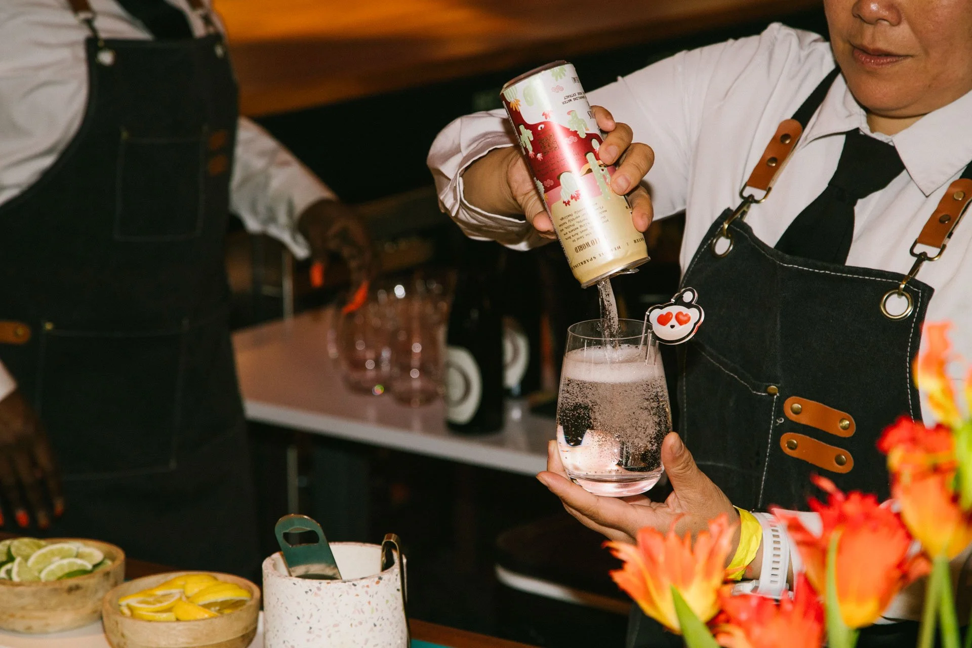 Bartender pouring a fizzy drink into a glass with decorative heart-shaped stir stick, behind a bar with bowls of sliced citrus fruits and colorful flowers.