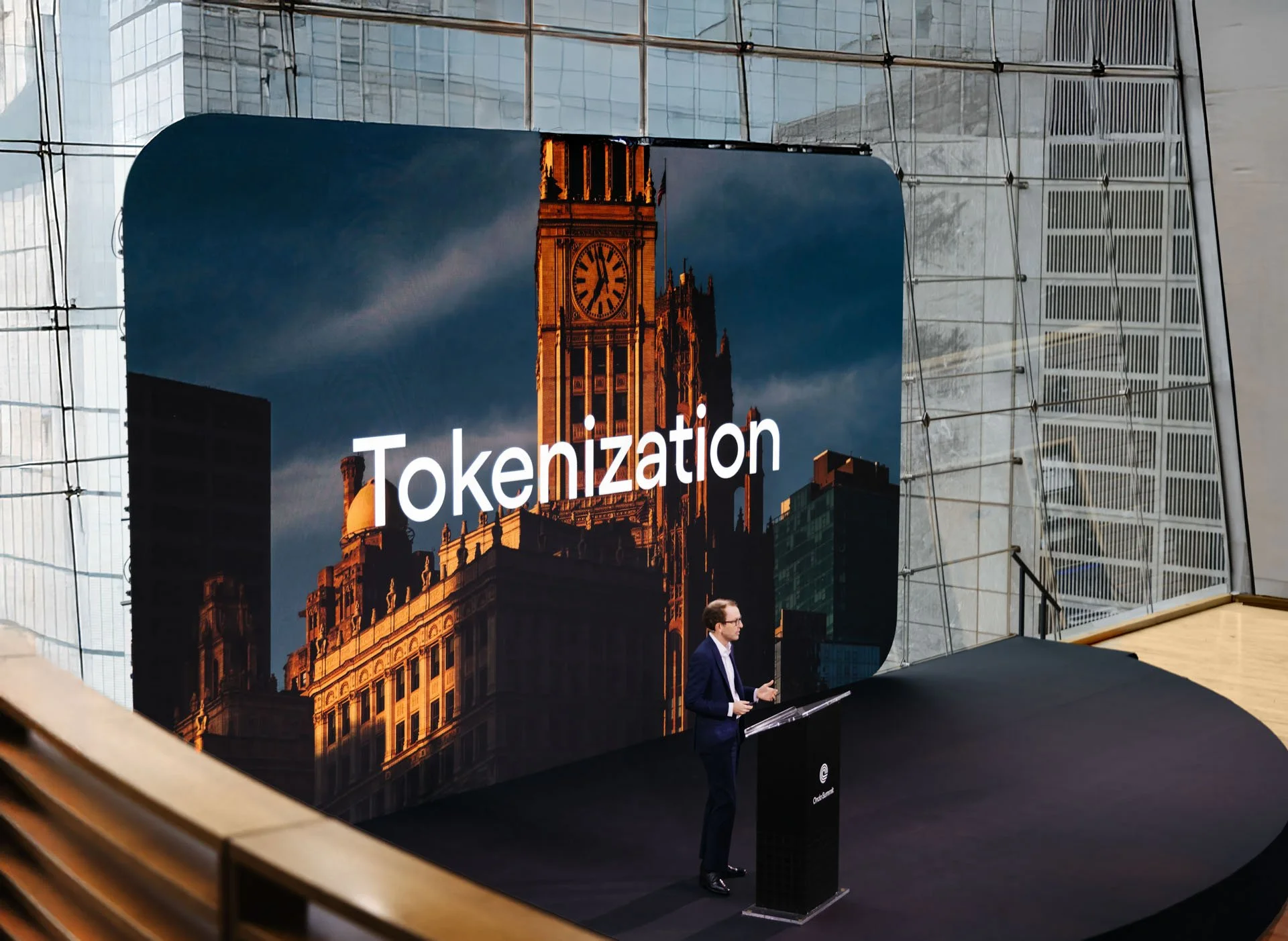 A man in a suit standing at a podium giving a presentation in front of a large screen displaying an image of Big Ben clock tower and the word 'Tokenization' in white text.