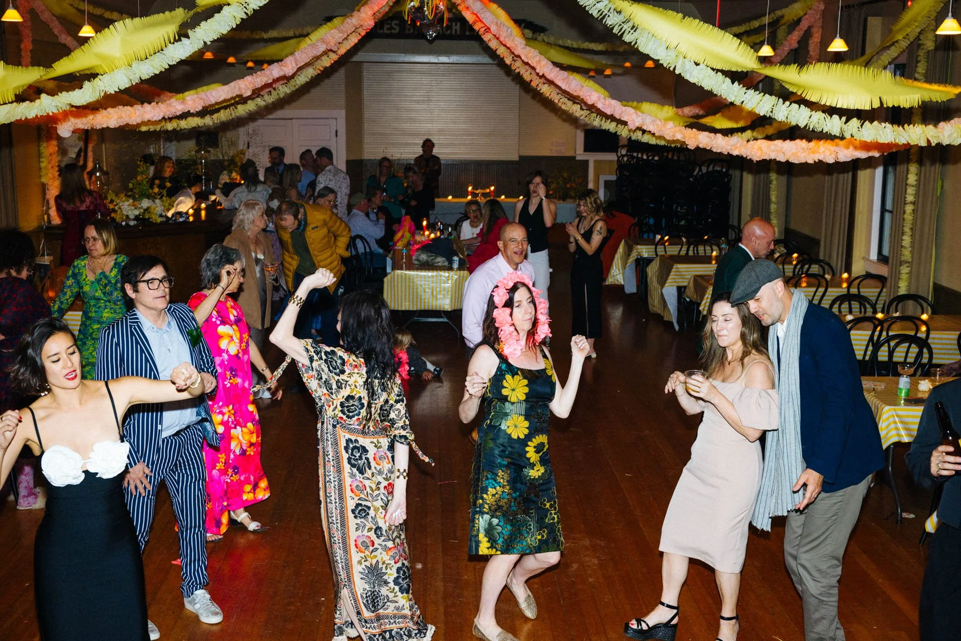 People dancing and socializing at a party with colorful paper streamers hanging from the ceiling, some wearing floral outfits and accessories, in a decorated venue with tables and chairs.