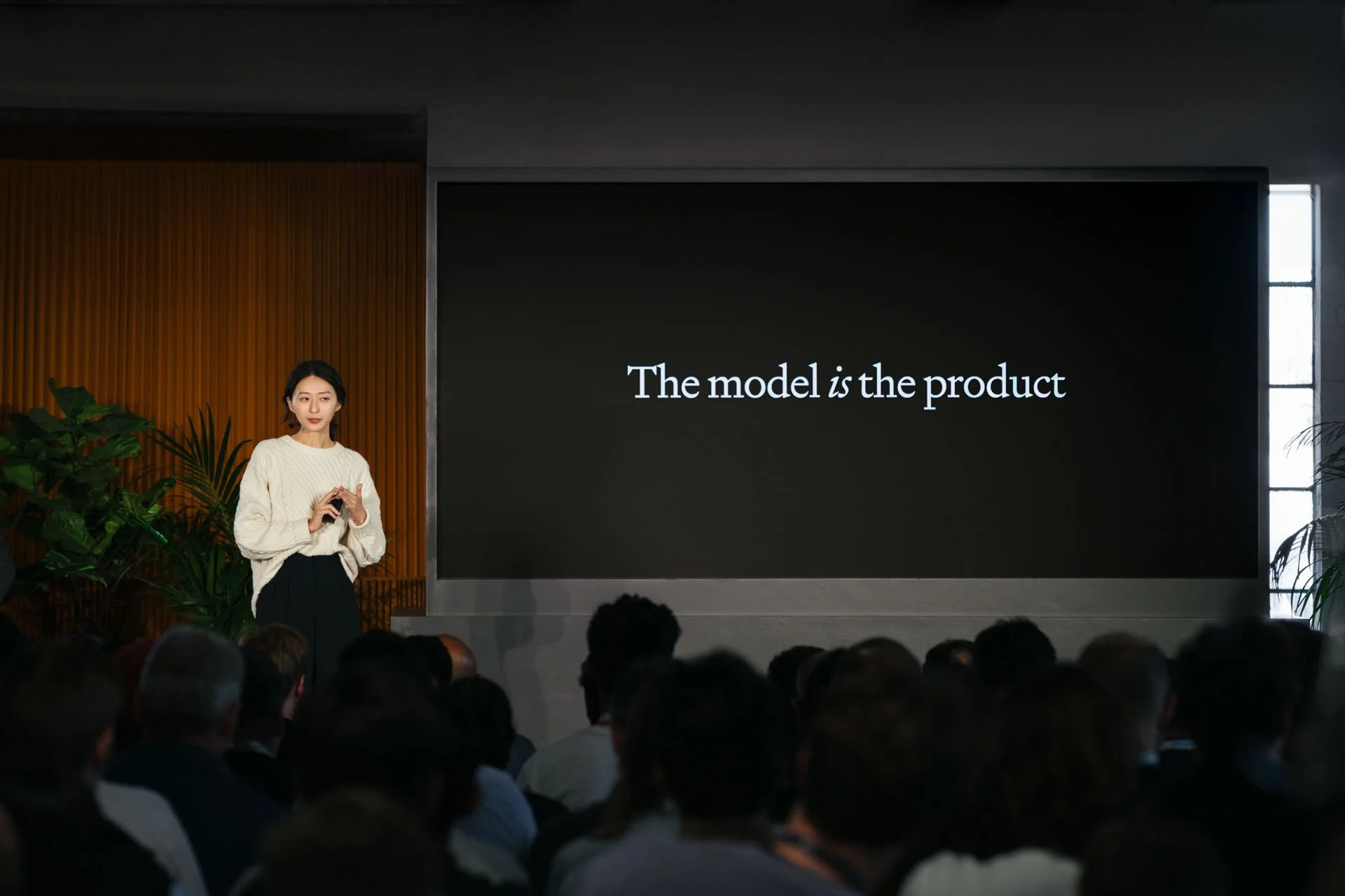 A woman giving a presentation in front of an audience with a large screen displaying the text 'The model is the product' in a modern conference room.