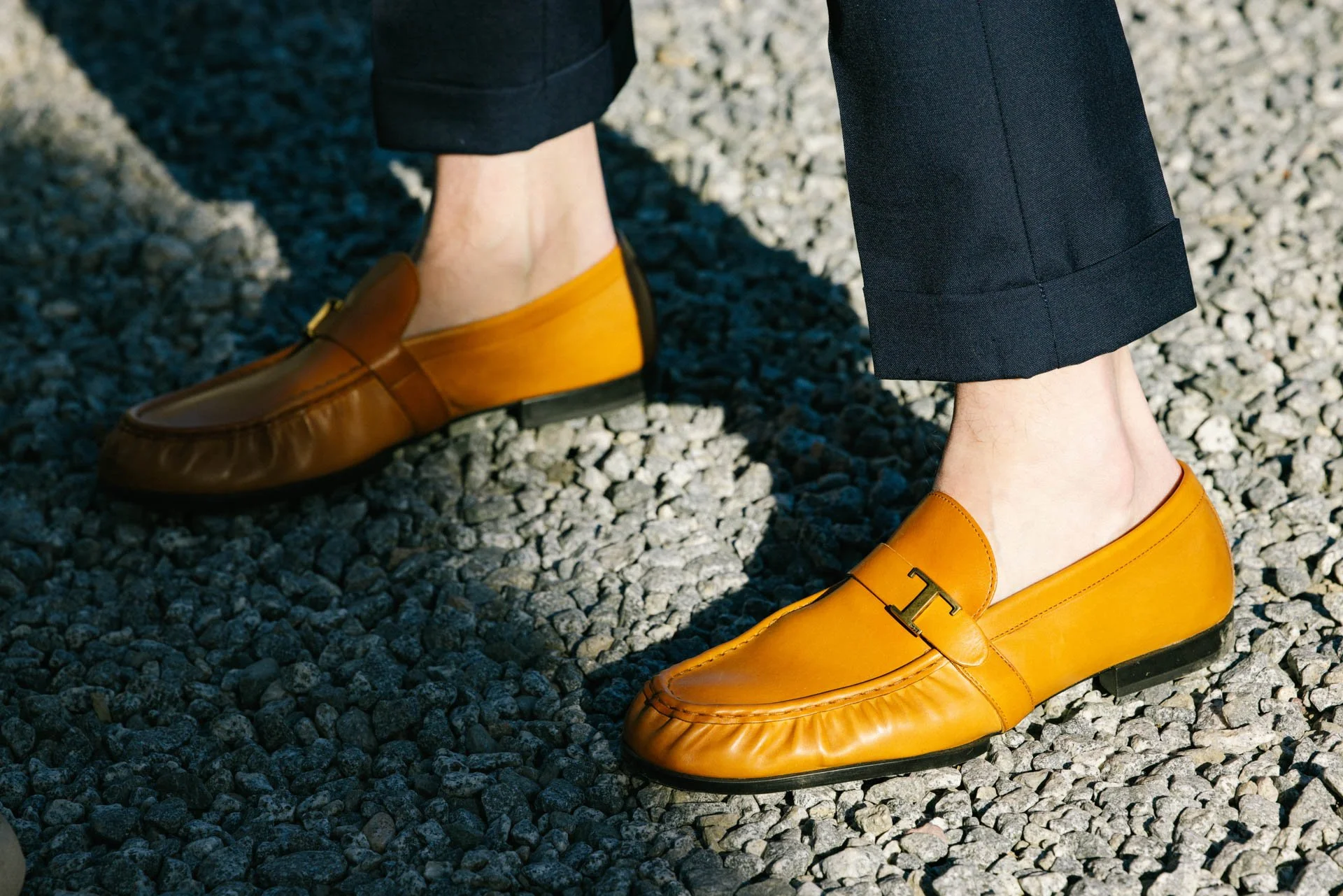 Person wearing black pants and yellow leather loafers with a metallic 'T' detail, standing on gravel surface.