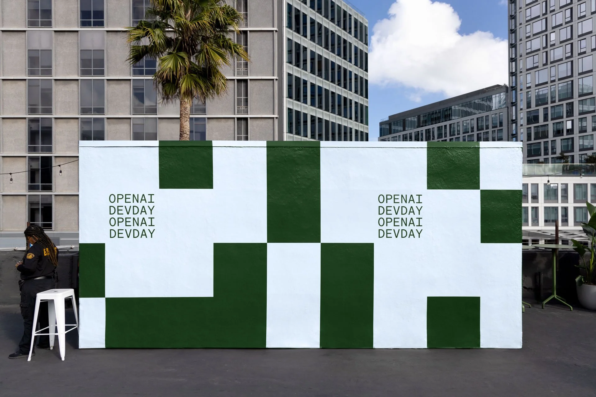 A white wall with green blocks and text that reads 'OPENAI DEV DAY' written twice on each side in an urban setting with tall buildings, a palm tree, and a person in a black jacket standing near a white chair.