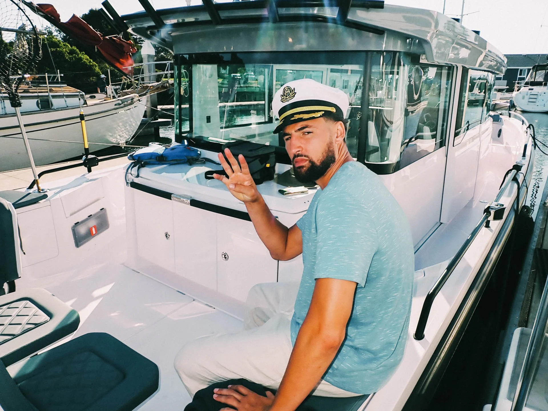 Klay Thompson for Cash App. A man dressed as a captain sitting on a white boat at a marina, making a peace sign with his hand. He is wearing a white captain's hat and a light blue t-shirt, with other boats visible in the background.