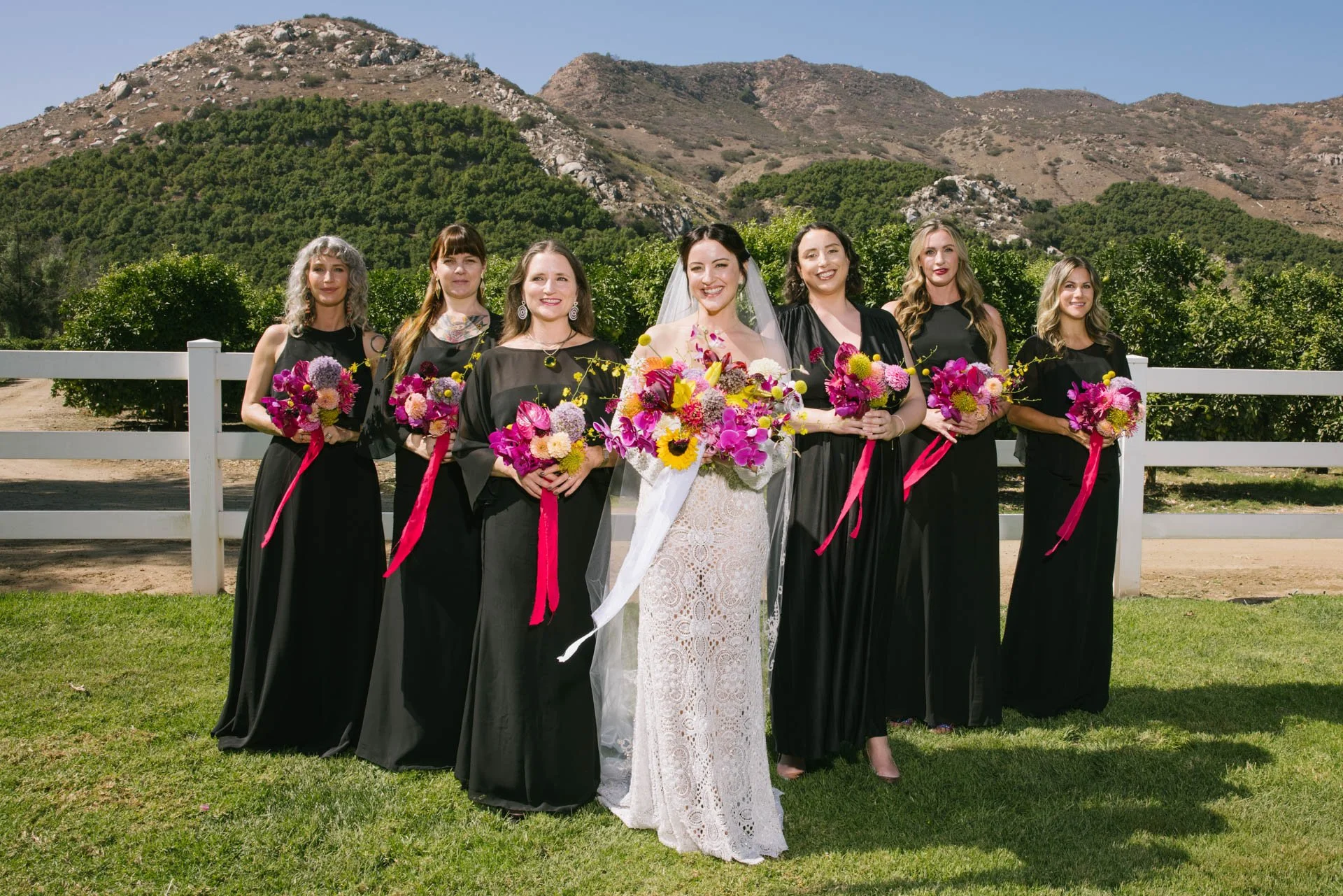 Bride in a white lace dress and six bridesmaids in black dresses standing outdoors with mountains in the background, holding colorful bouquets of flowers.