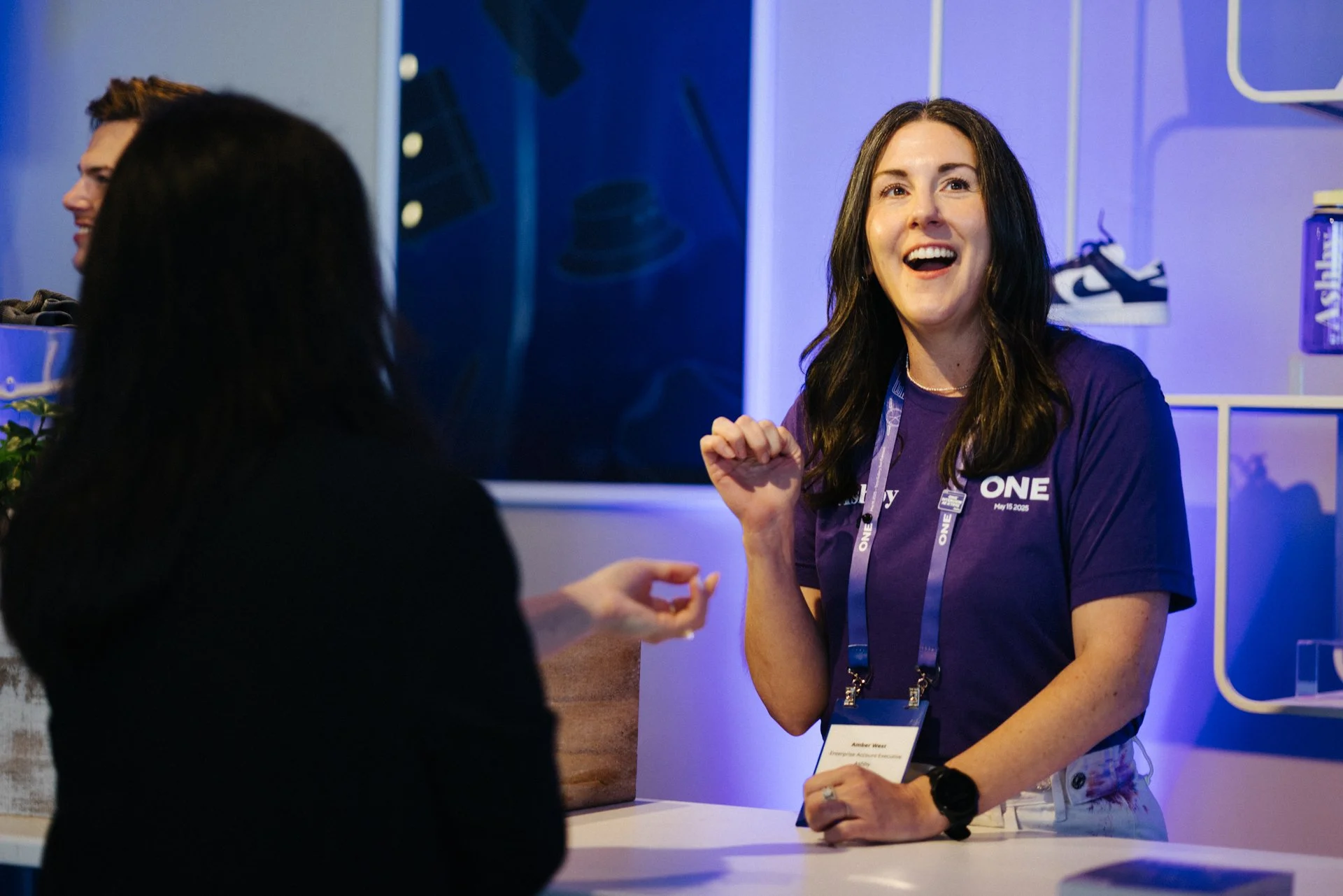 A woman with long dark hair, wearing a purple shirt with conference lanyards, is smiling and engaging in conversation with a person whose back is facing the camera. The background appears to be a booth or exhibit space.