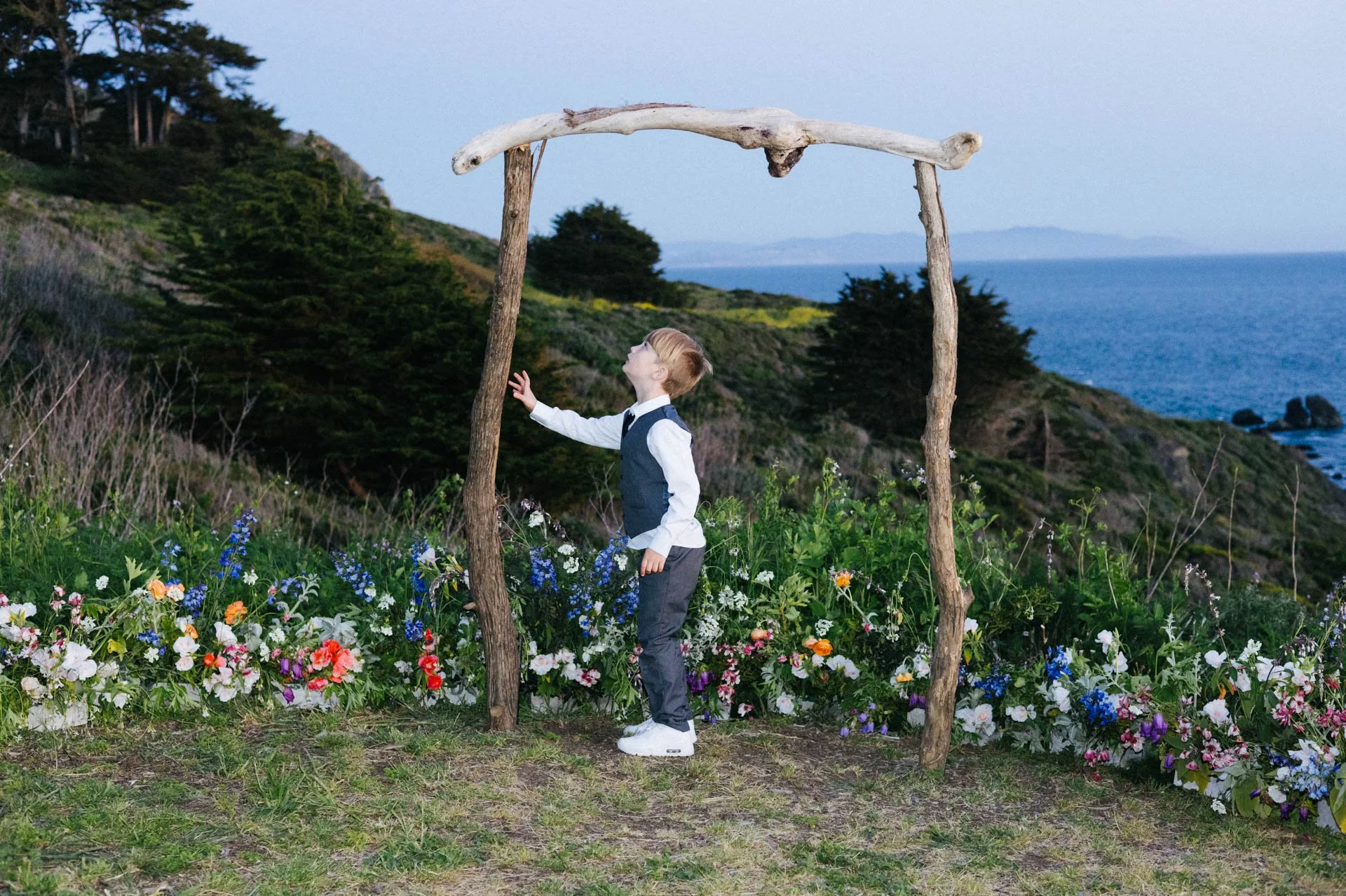 A young boy in a white shirt, gray vest, and dark pants stands under a rustic wooden arch made of driftwood on a grassy and flowered field, with ocean and hillside in the background, gazing upward.