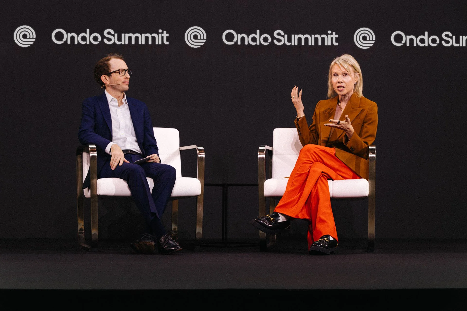 Two individuals seated on stage at the Onde Summit, engaged in conversation. The man on the left is dressed in a navy suit with glasses, and the woman on the right is wearing a brown blazer and bright orange pants, gesturing as she speaks. The black 