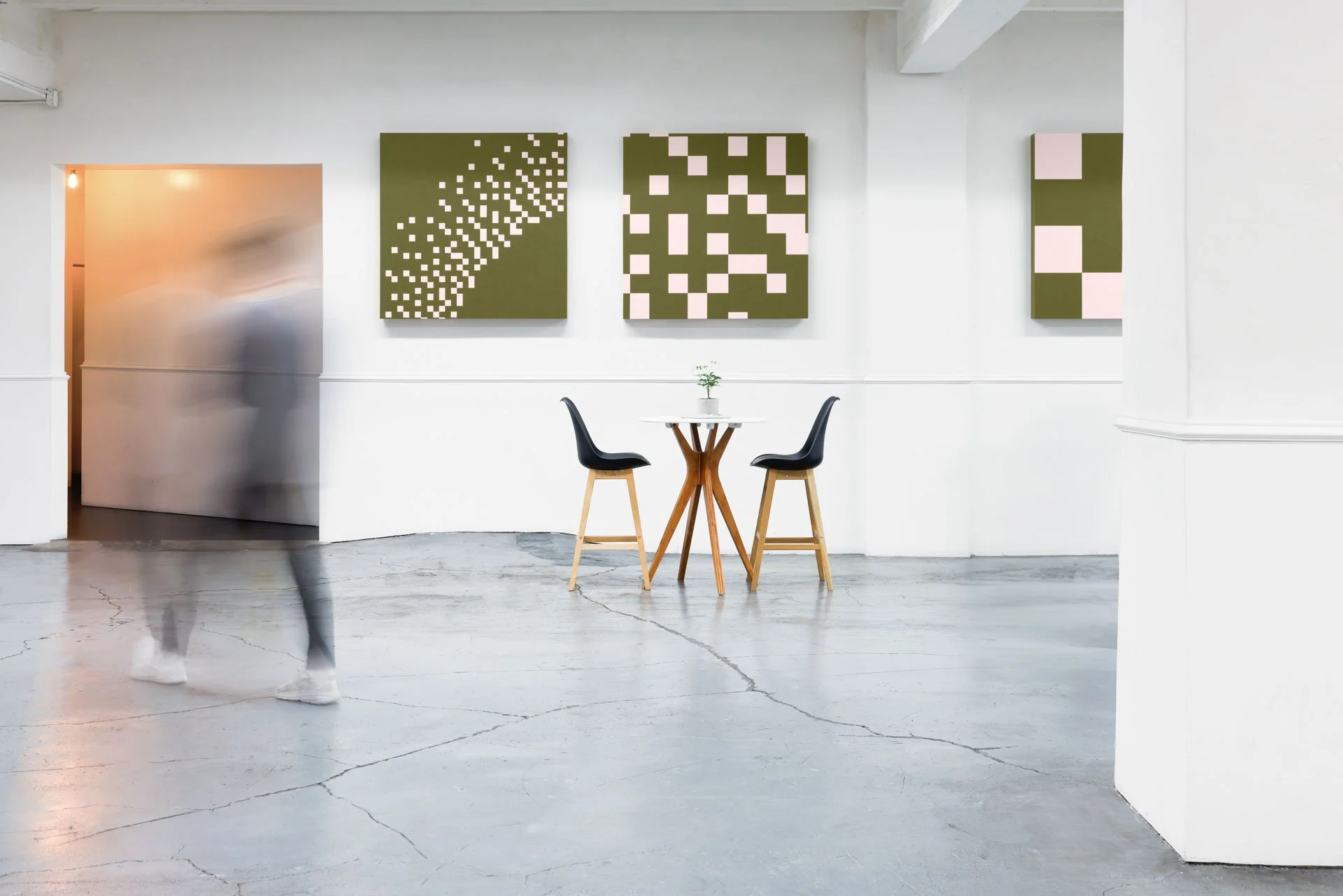 An art gallery with four geometric paintings on the white wall, a small round table with a plant and two black chairs. A woman walks past, slightly blurred, near the entrance to a hallway.