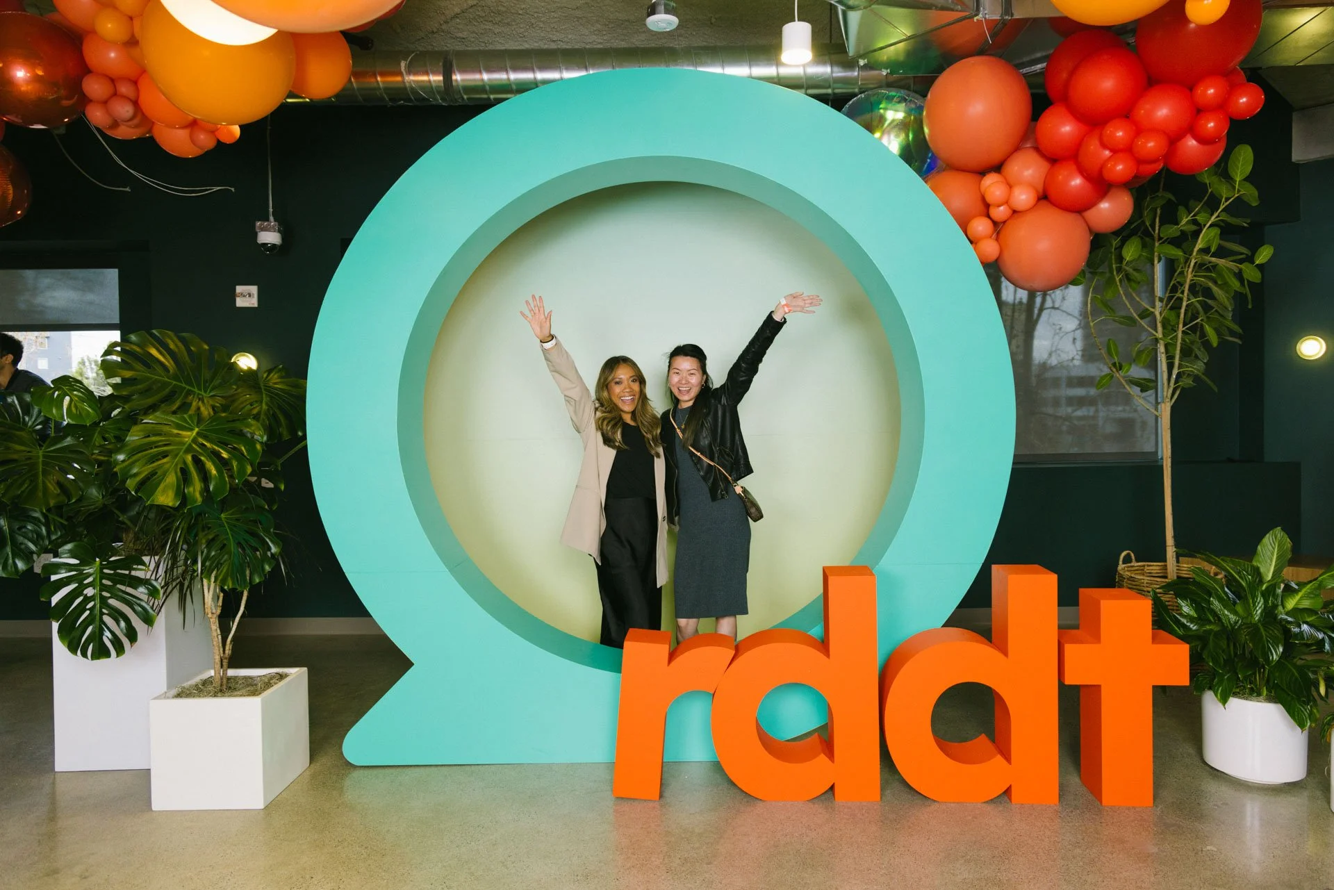 Two women standing inside a large, teal-colored question mark sculpture, smiling, with the word 'rddt' in large orange letters at their feet, in an indoor space decorated with plants and balloons.