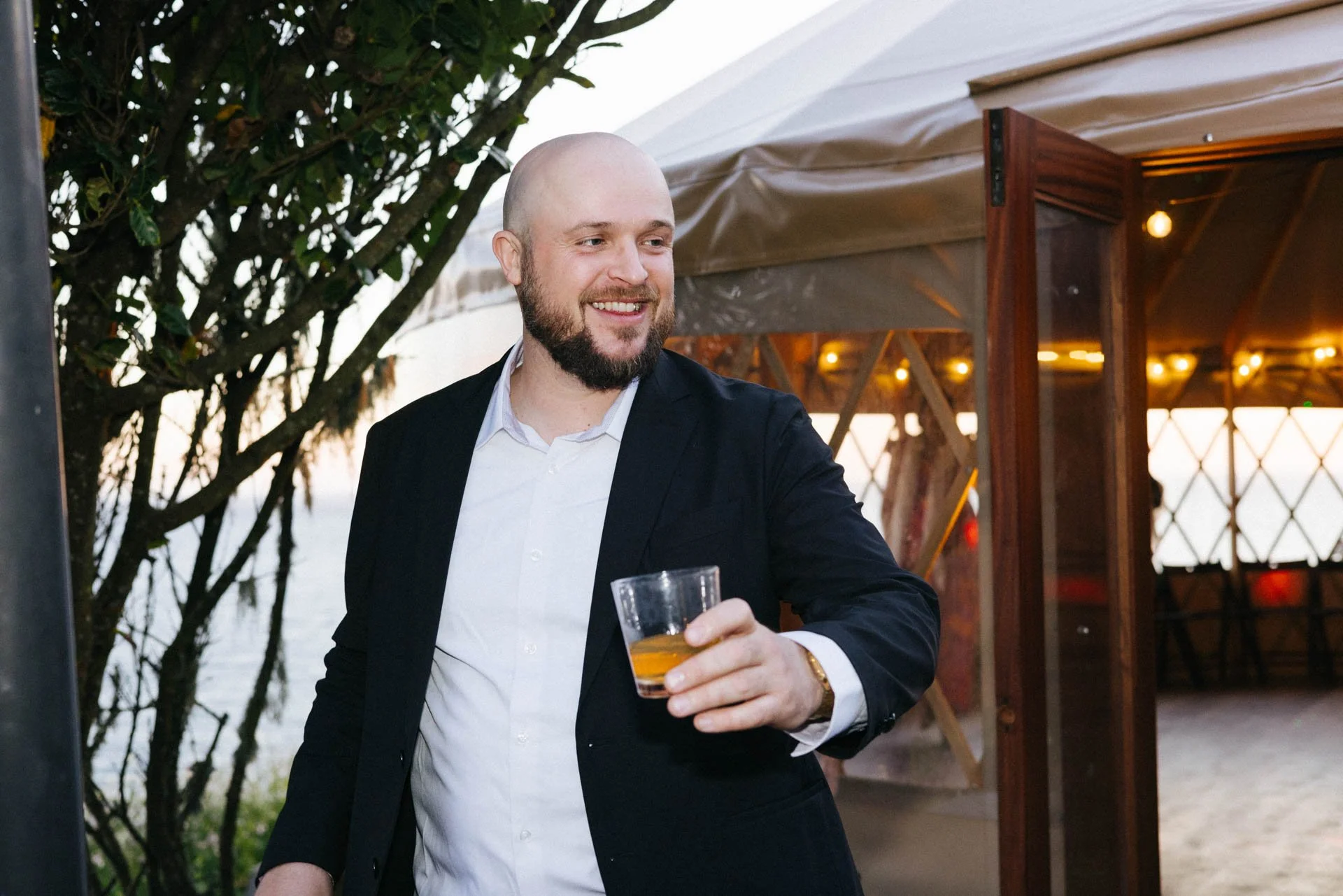A man with a beard and bald head smiling while holding a glass of drink at an outdoor event near water and a tent with string lights.
