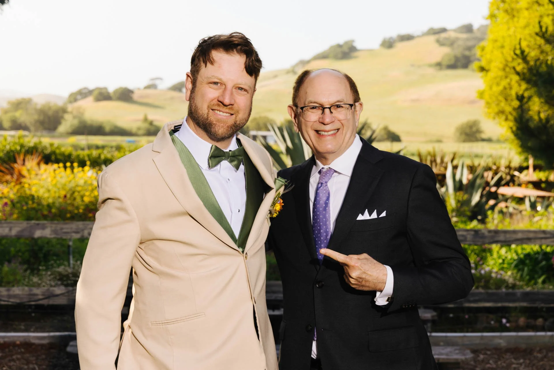 Two men dressed in formal wedding attire standing outdoors with a lush landscape in the background, smiling at the camera. The man on the left wears a beige suit with a green bow tie, and the man on the right wears a dark suit with a purple tie, poin