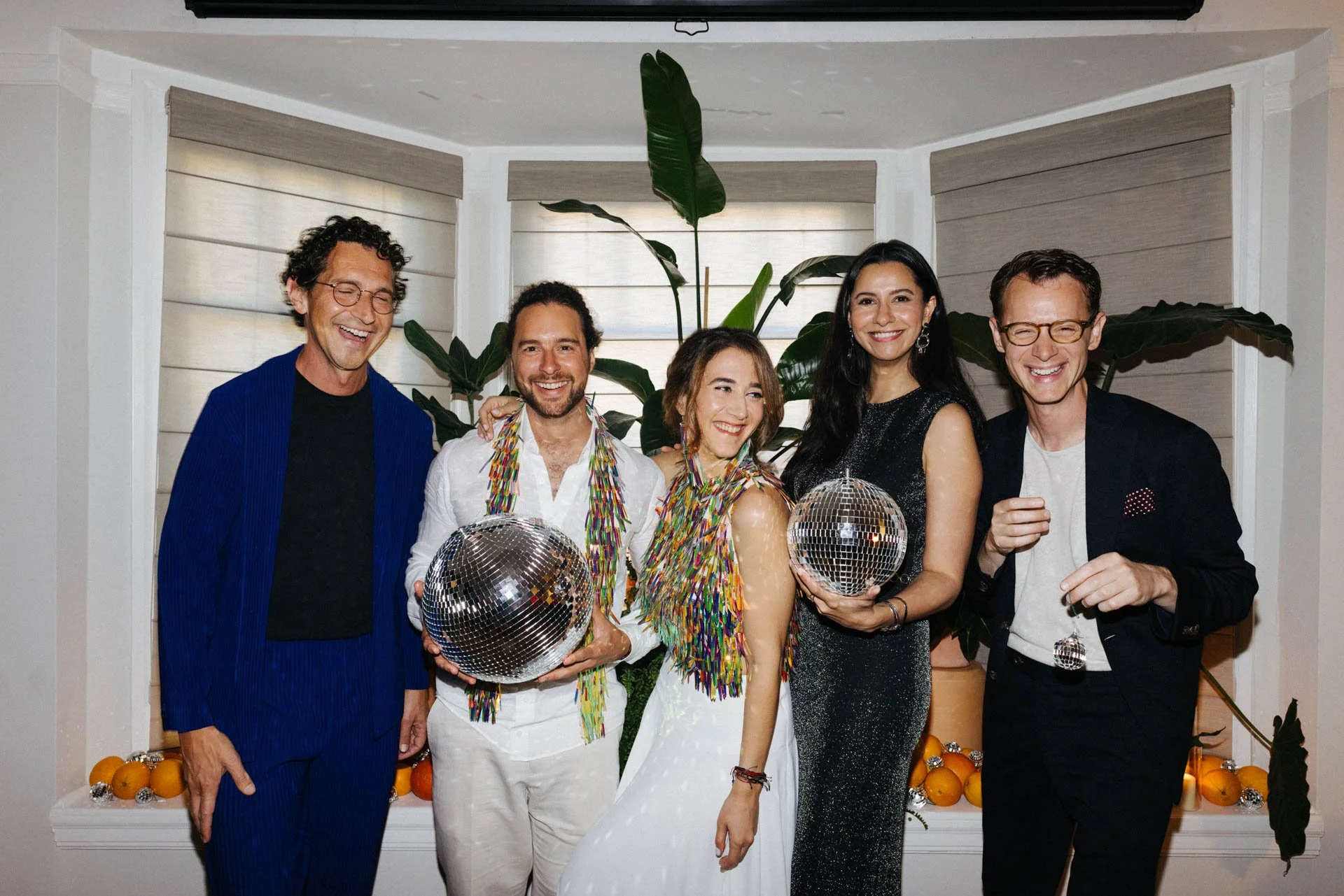 Group of five people celebrating, holding disco balls, with a backdrop of green foliage and oranges