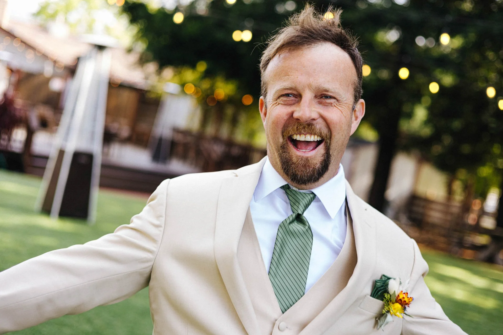 A smiling man in a cream suit jacket and green striped tie, standing outdoors in a park or garden, with trees and string lights in the background.
