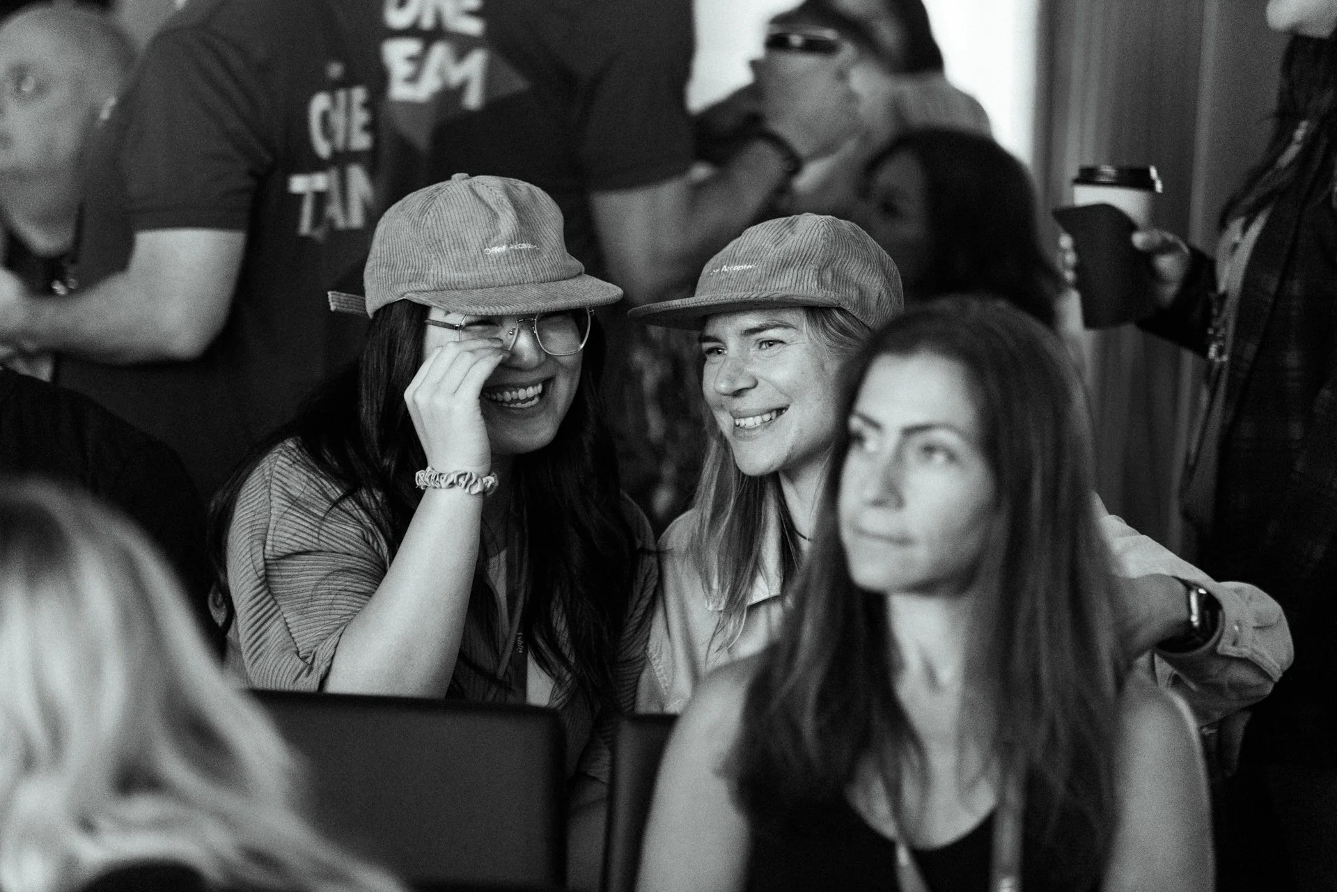 Three women in a social gathering smiling and enjoying each other's company, two wearing hats and the woman in the foreground has long hair and is wearing a sleeveless top.