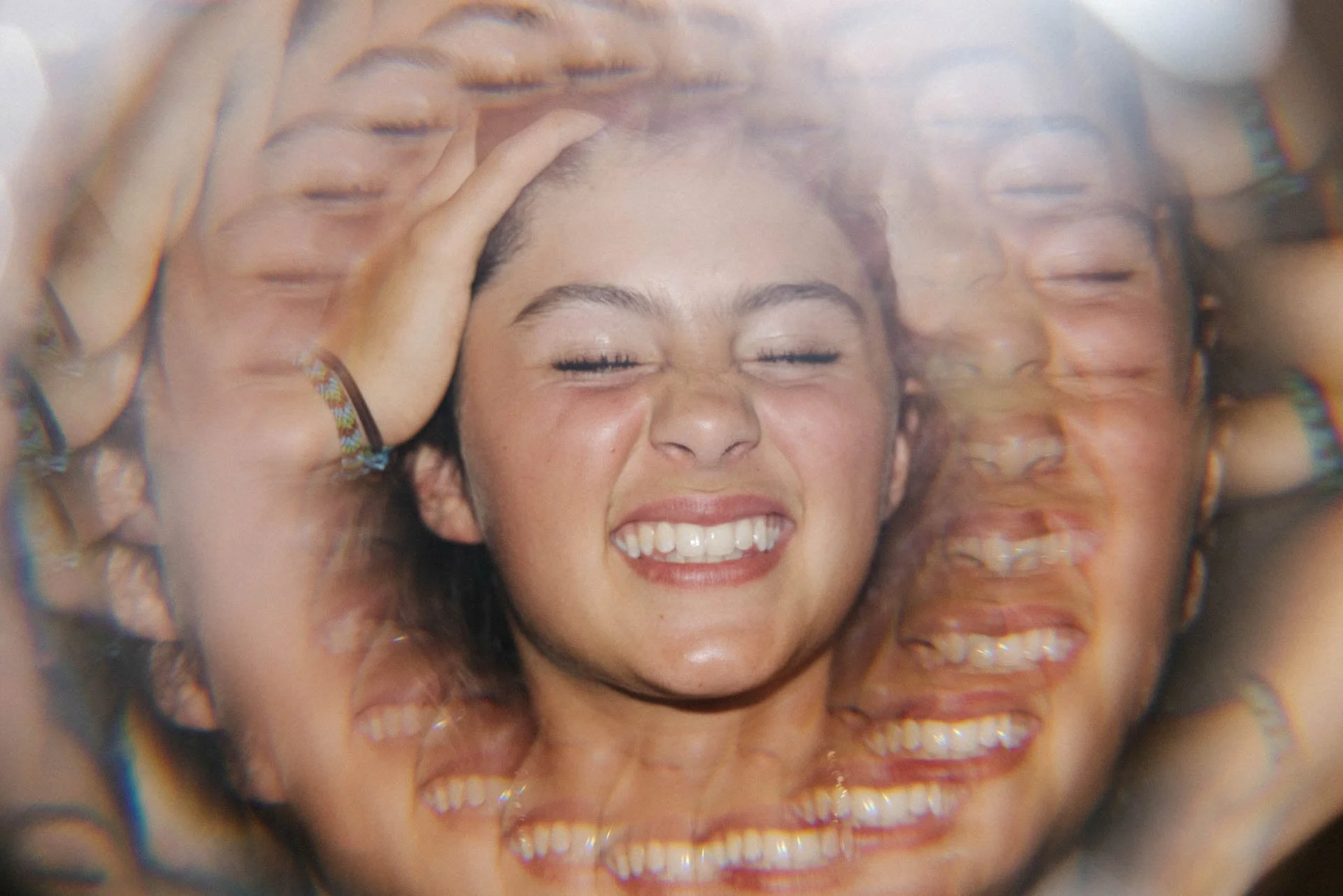 A person with curly hair smiling with eyes closed, surrounded by multiple overlapping images of the same person, creating a kaleidoscope effect.