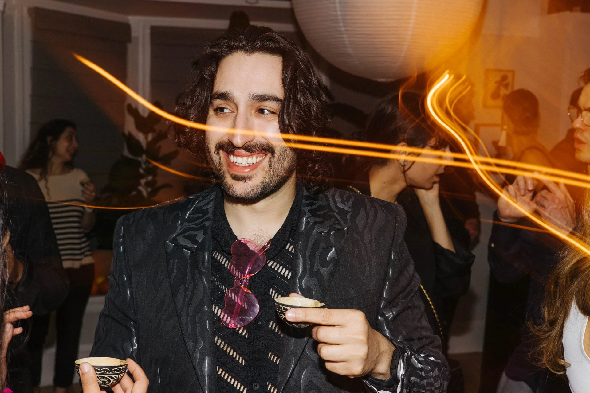 A man with curly hair and a beard smiling at a party, holding a drink, with colorful light streaks and other people in the background.