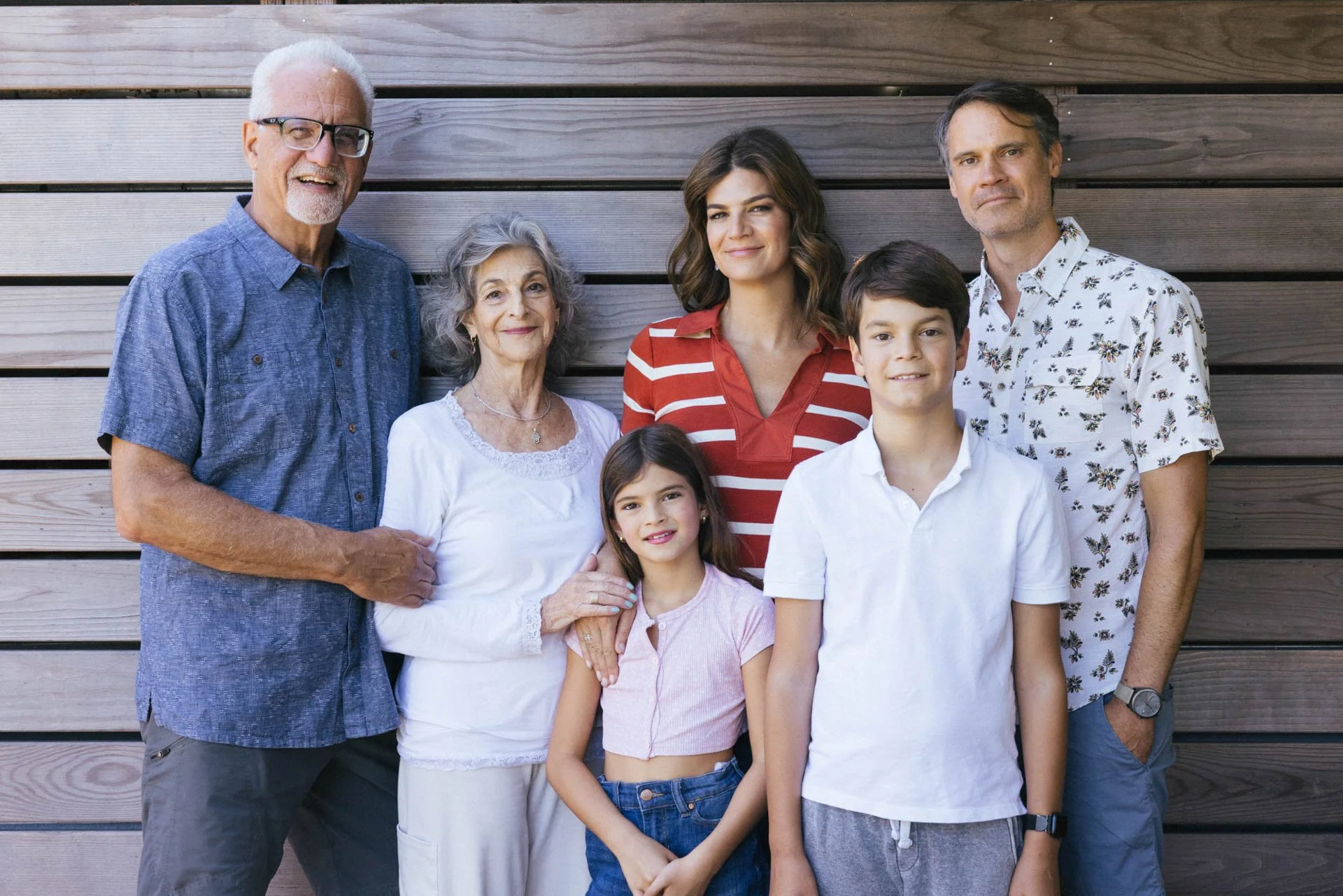 Multi-generational family standing outdoors against a wooden wall, smiling at the camera.