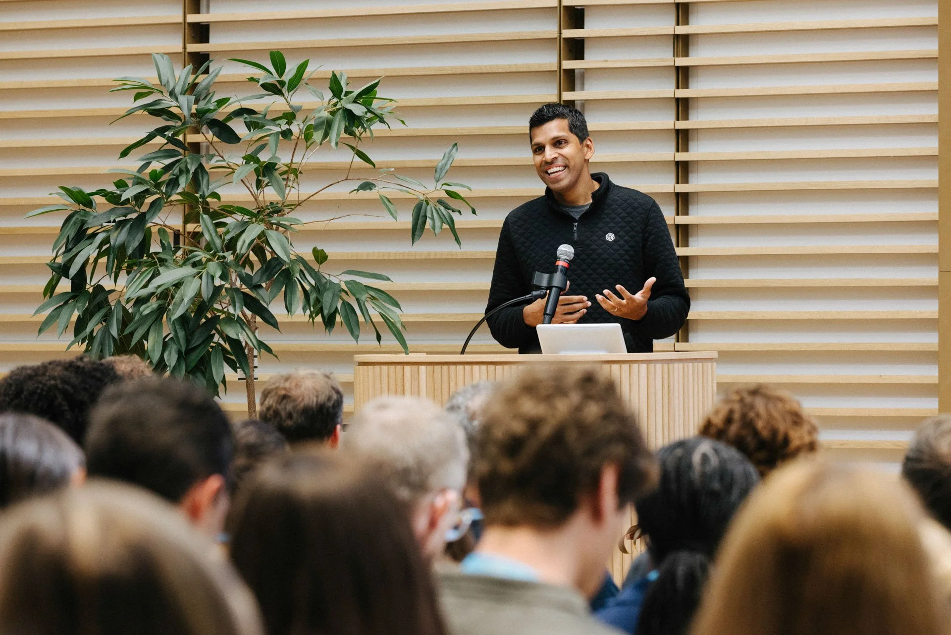 A man speaking at a podium with a microphone, smiling, in front of a group of people, with a large green plant and wooden slatted wall in the background.