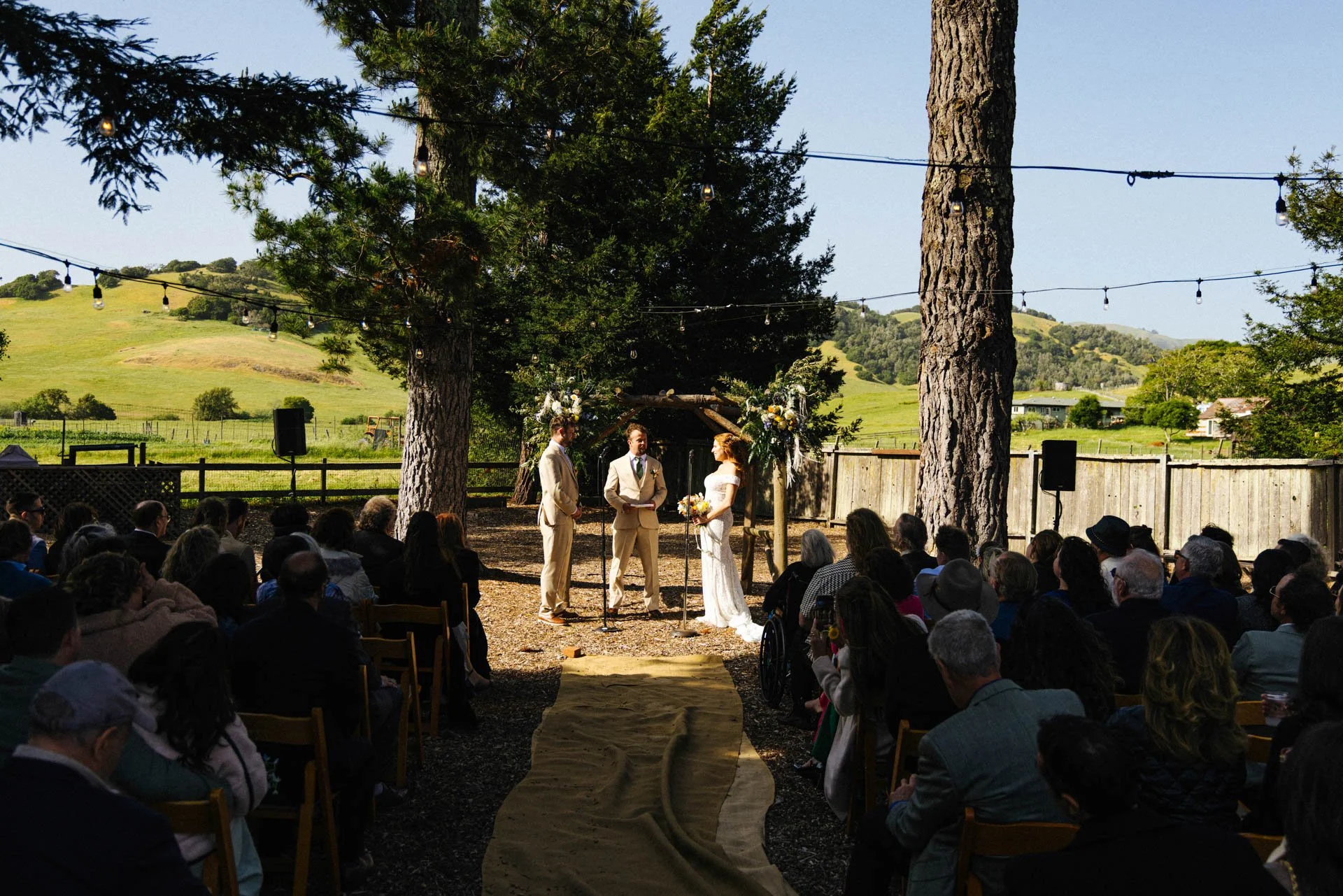Outdoor wedding ceremony taking place beneath tall trees with string lights. The couple stands at the altar with an officiant, surrounded by guests seated in chairs on either side, with scenic green hills in the background.