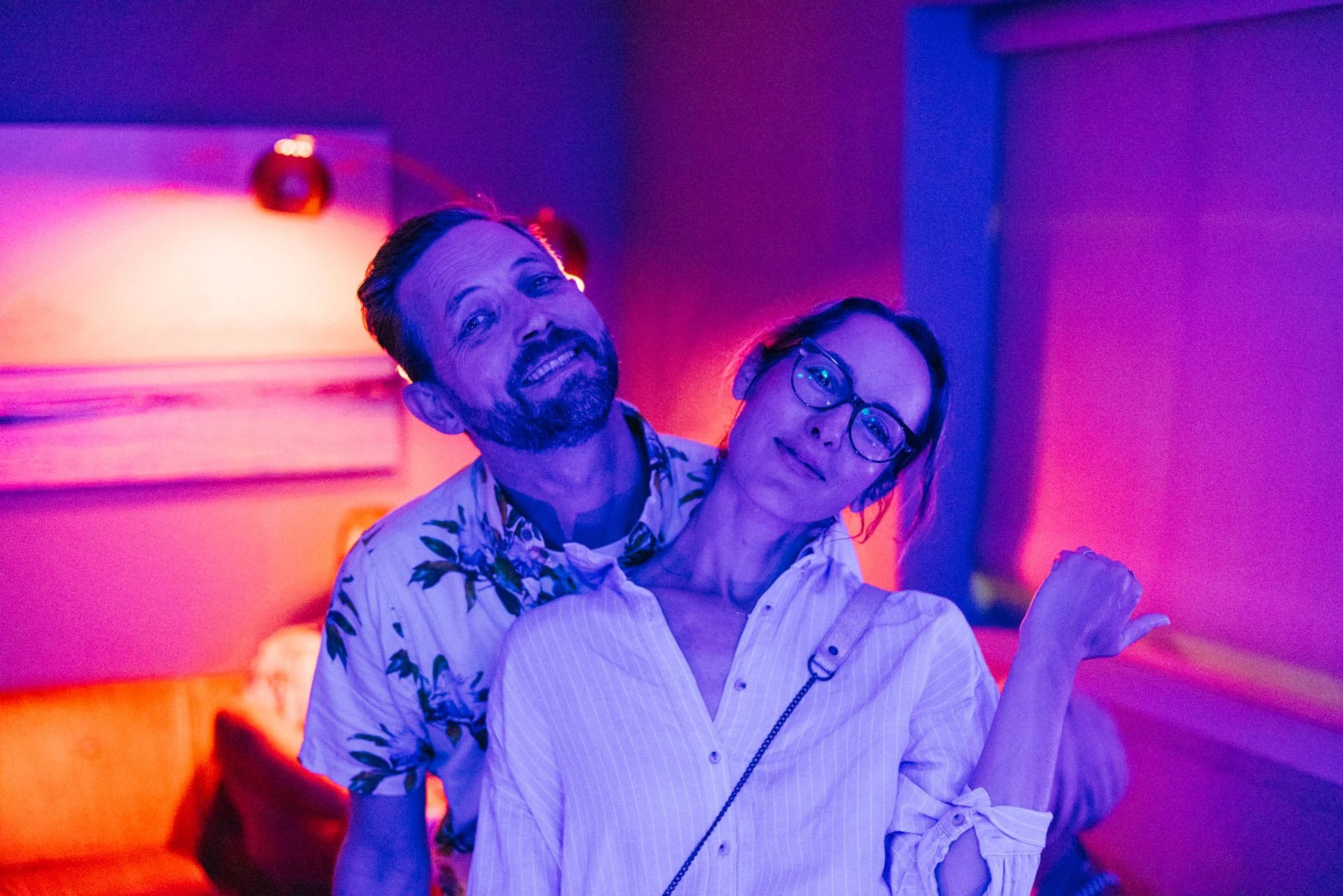 A man and woman with glasses in a dimly lit room with neon pink and blue lights, posing together.