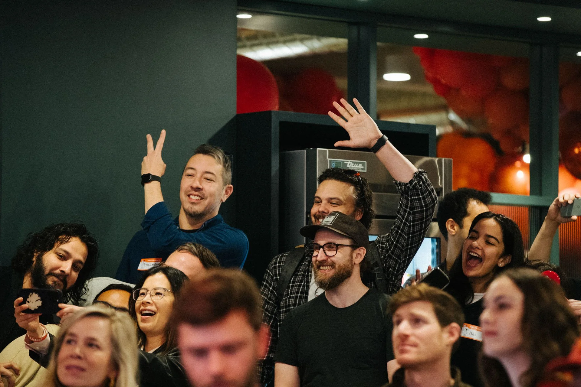 Group of people at an event, some smiling, some taking photos, one person raising their hand, in a lively indoor setting.