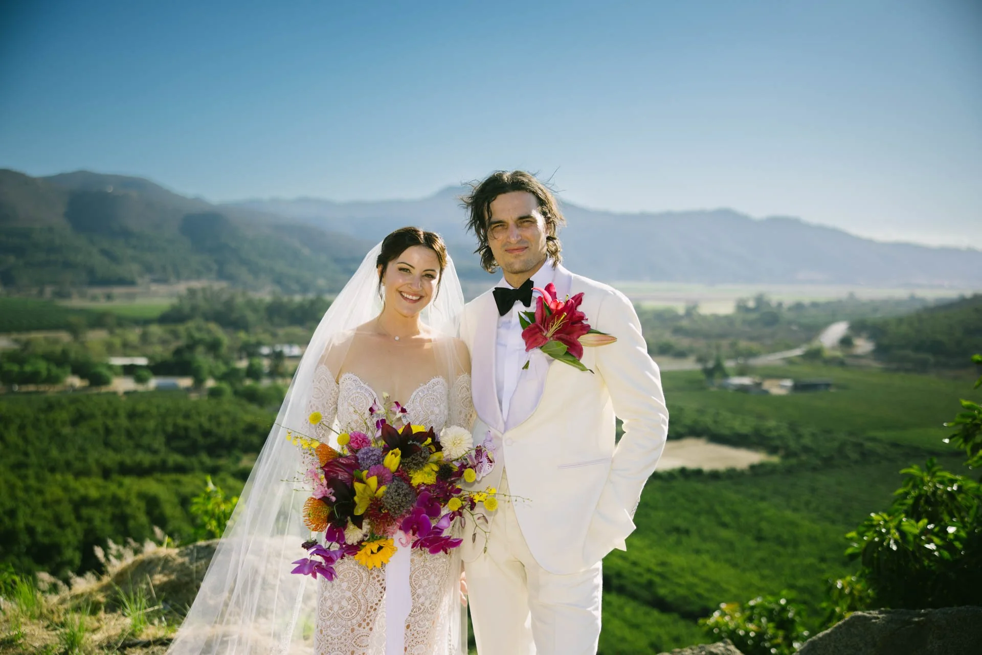 A bride and groom standing together outdoors on a sunny day, with a scenic view of green fields and hills in the background. The bride is holding a colorful bouquet of flowers, and the groom is holding a pink flower on his lapel. Both are dressed in 