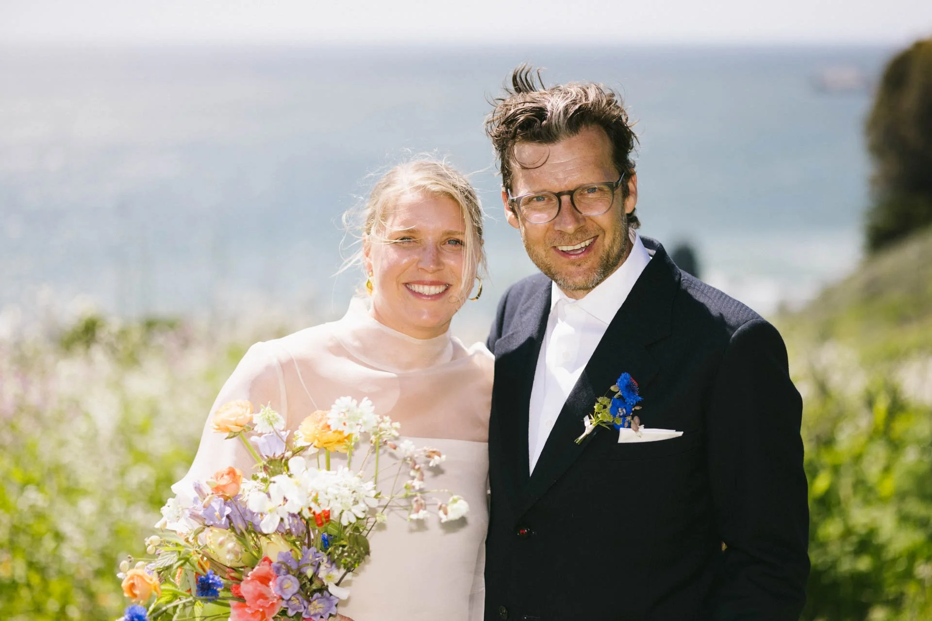 A smiling bride in a white wedding dress holding a colorful bouquet, standing next to a smiling groom in a black tuxedo with a blue flower boutonniere, outdoors with a scenic ocean view in the background.