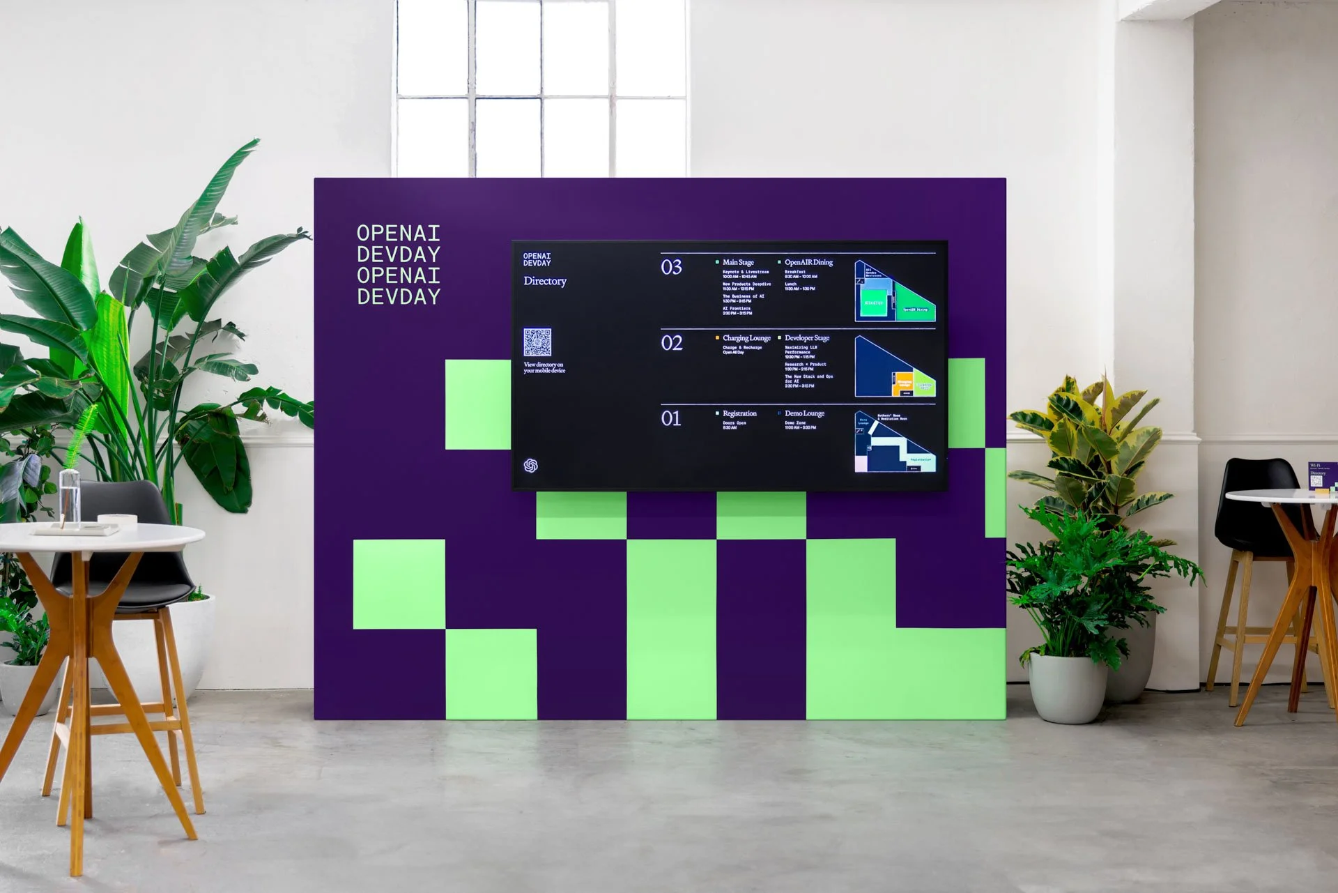 Digital displayboard with directions at a conference or event in a modern space, surrounded by plants and small tables.