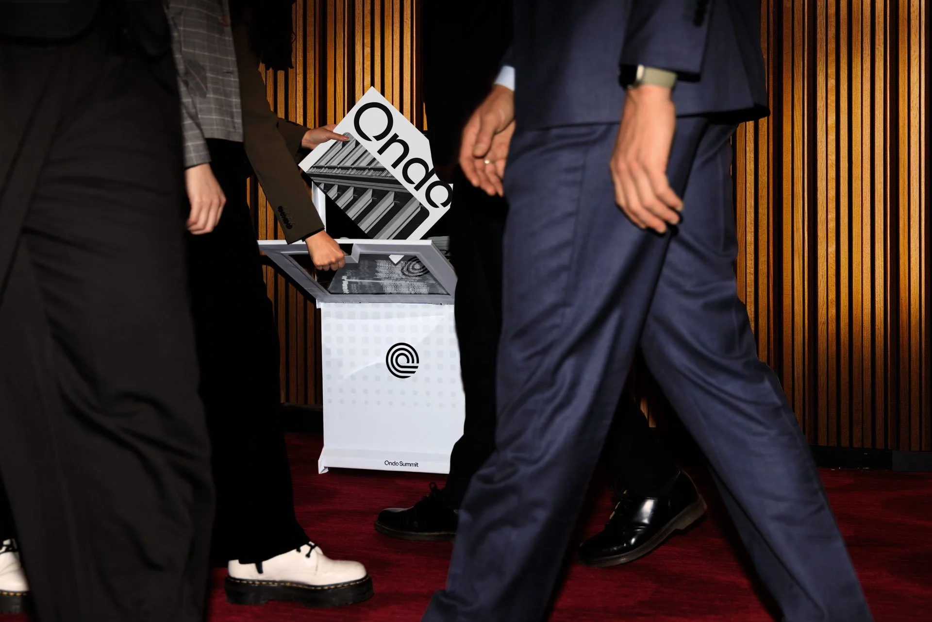 People in business attire surrounding a white podium with a black logo, with one person placing a ballot into the podium at the Ondo Summit in a room with wooden paneling.