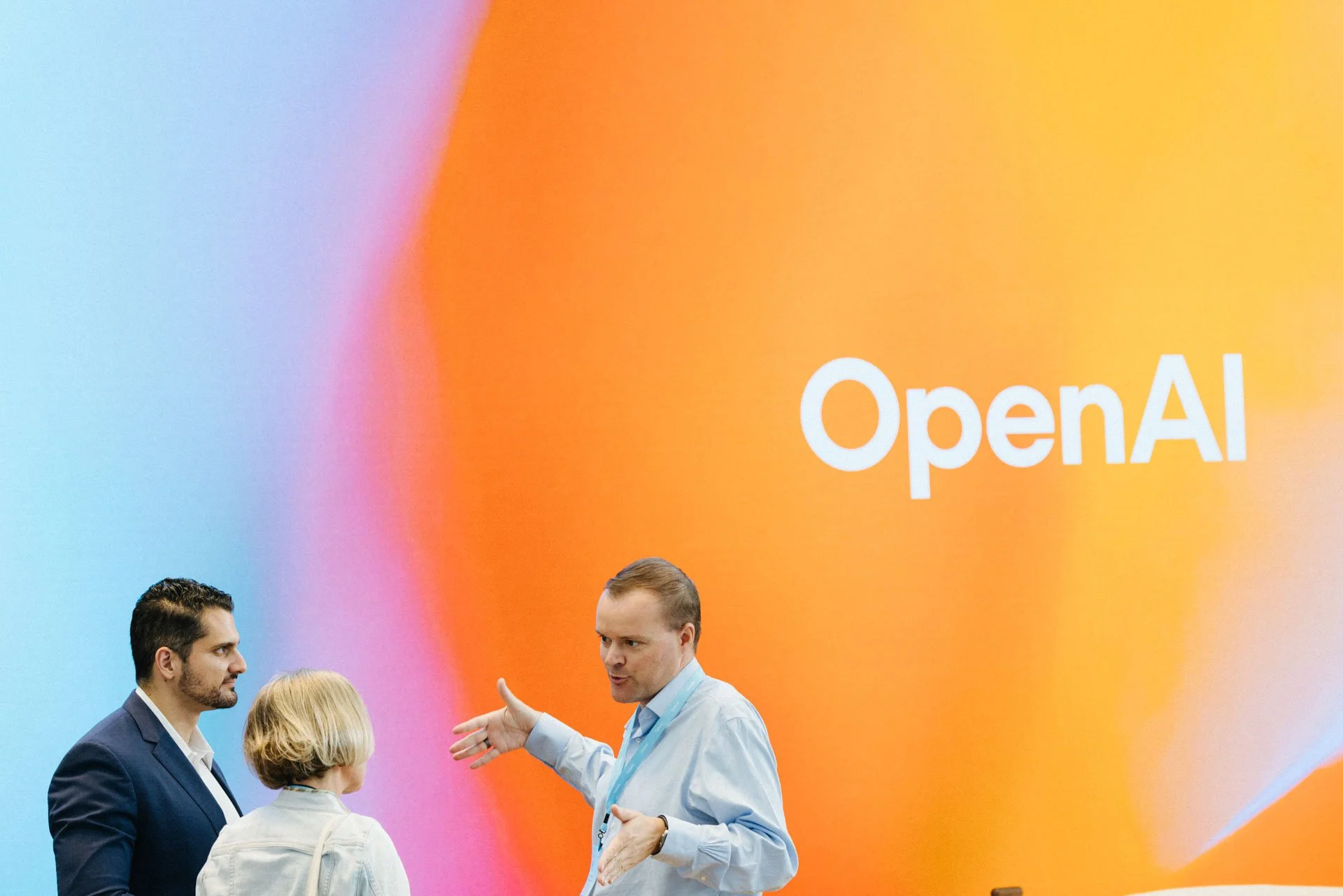 Three people having a discussion in front of a large screen with the OpenAI logo.