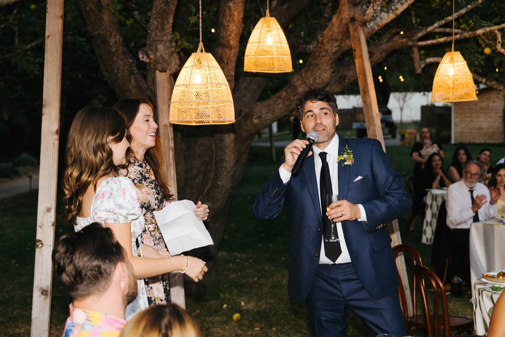 A man dressed in a blue suit gives a speech with a microphone at an outdoor celebration or wedding reception. Two women are listening warmly, with one holding a piece of paper. Guests are seated at tables in the background under string lights and han