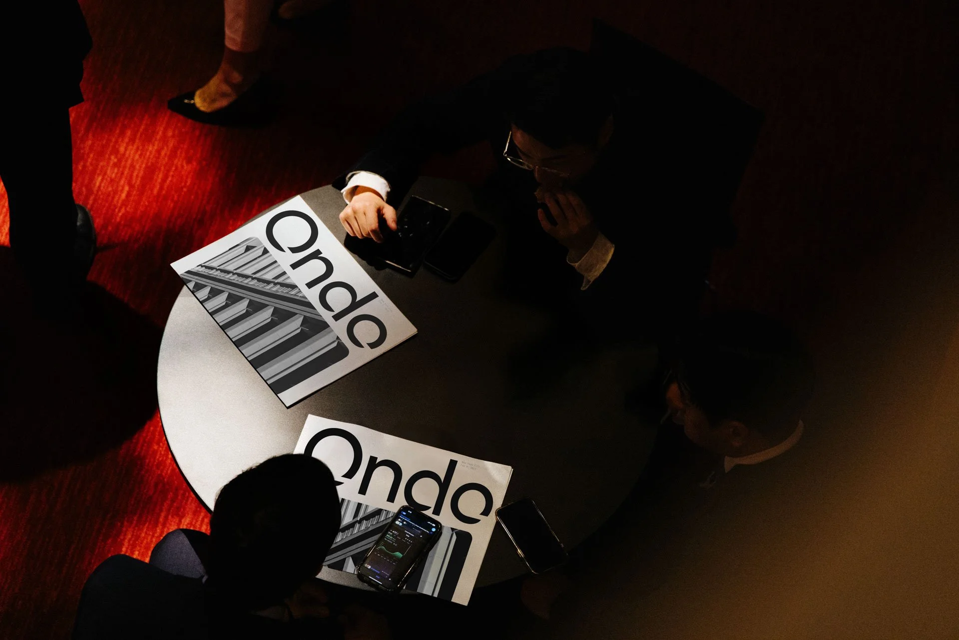 Three people are gathered around a round table with election campaign signs that say 'Undec' with a black and white building image on them. One person is sitting, and two are standing, all engaged with their phones.
