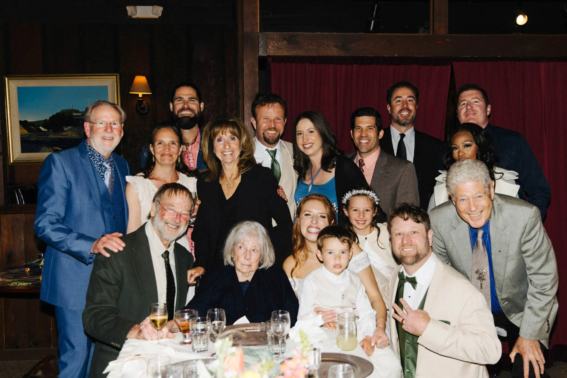A large group of people celebrating at a dinner party, dressed in formal and semi-formal attire, gathered around a table with glasses and a floral centerpiece, indoors with wooden walls and red curtains.