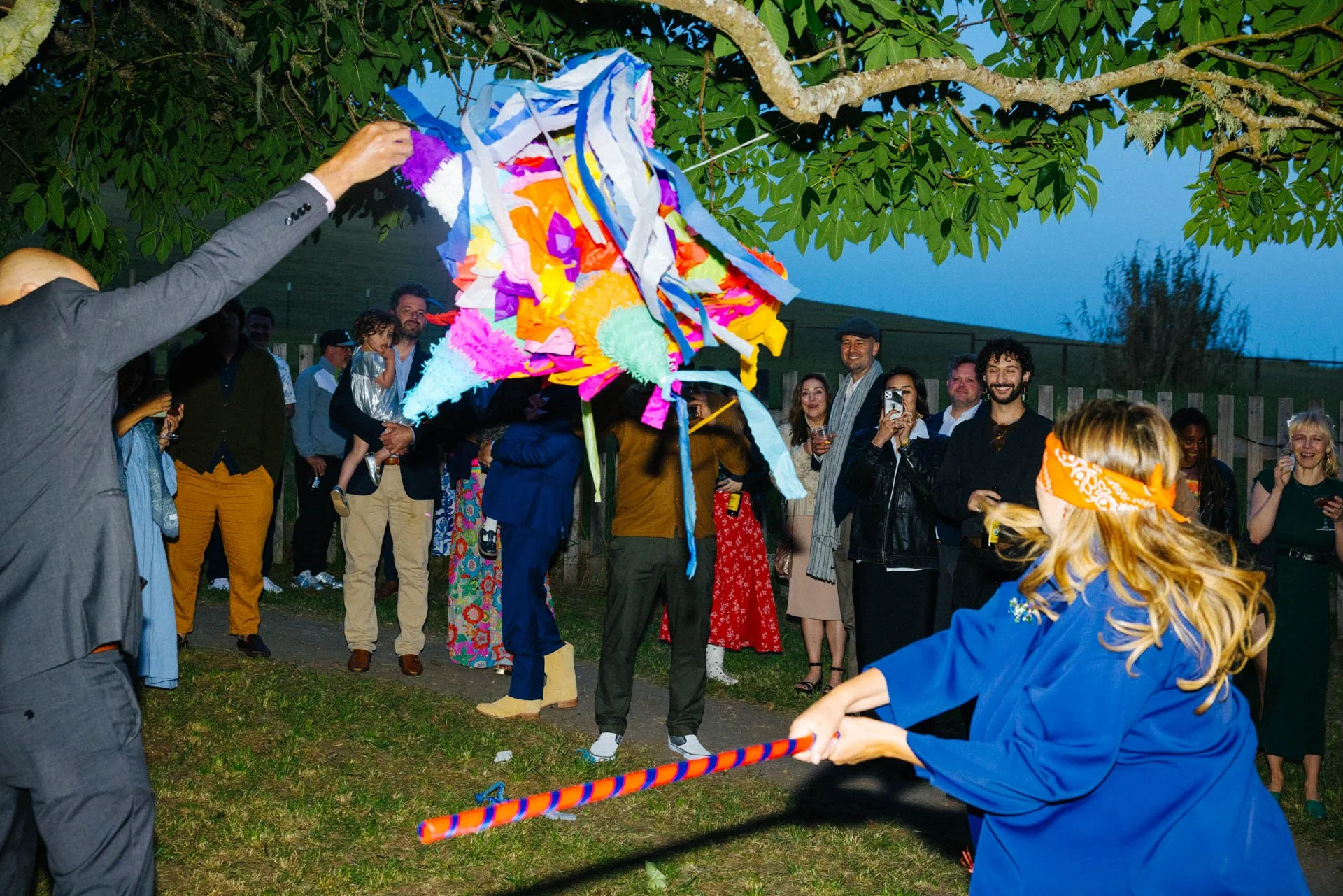 People celebrating outdoors during dusk, with some playing a piñata game using a colorful stick, surrounded by others watching and smiling.