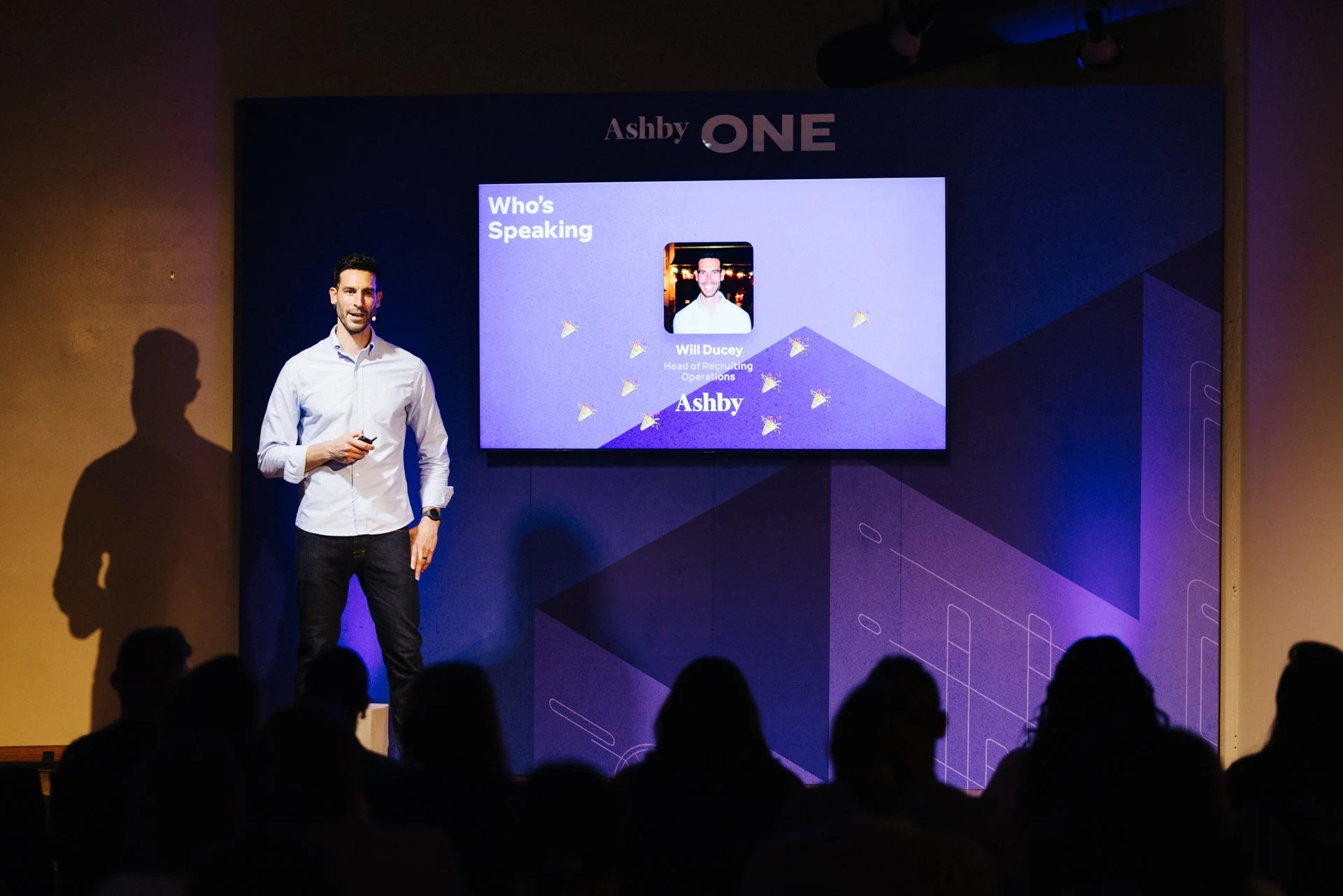 A man standing on a stage next to a large screen displaying a presentation slide with the title "Who's Speaking" and a photo of Will Ducey, Head of Recruiting Operations at Ashby. The man is holding a remote and is dressed in a white shirt and dark p