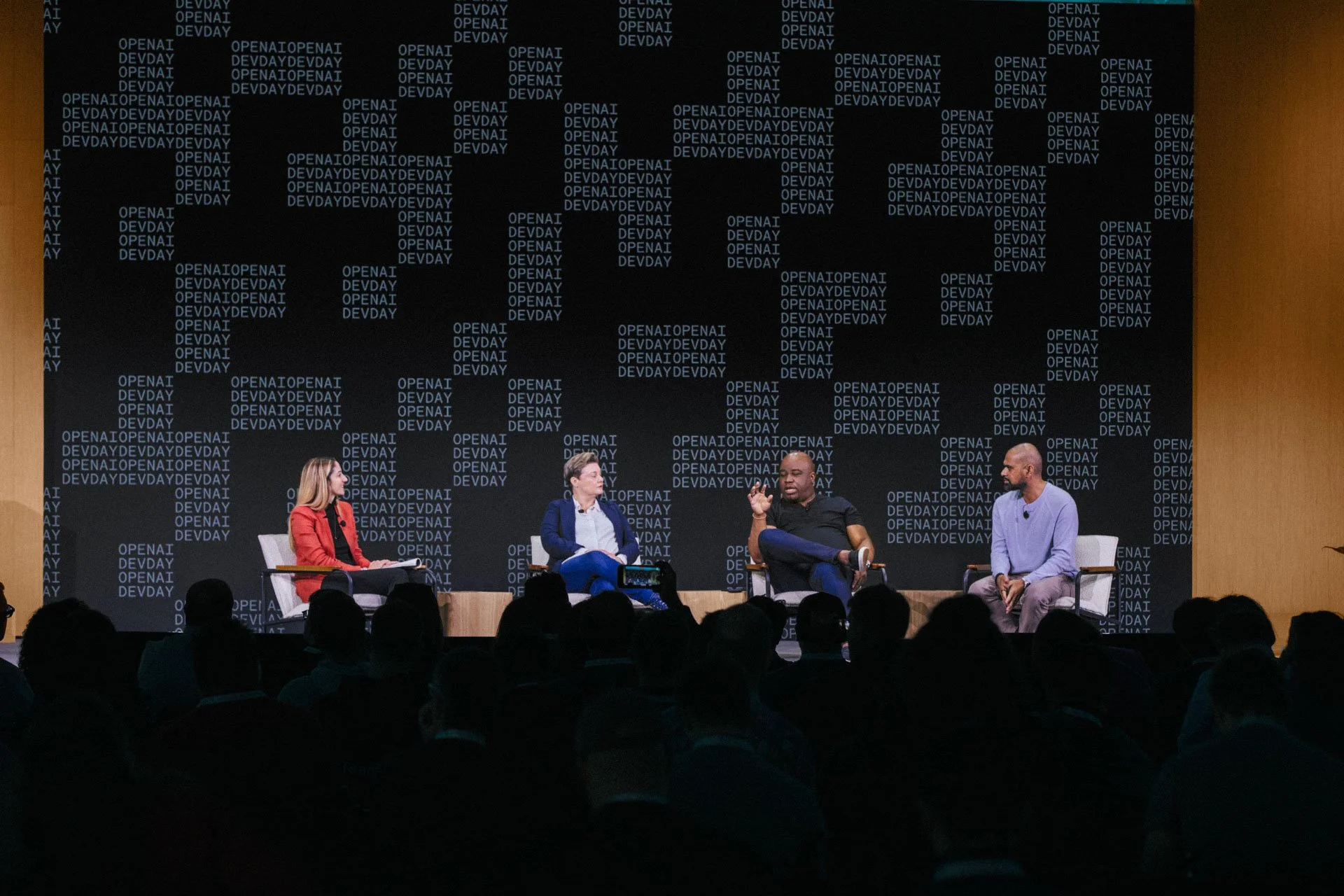 Four people seated on stage in front of a dark background with the words 'OPENAI' and 'DEVDAY' repeated, participating in a panel discussion at a conference or event. An audience is visible in the foreground.