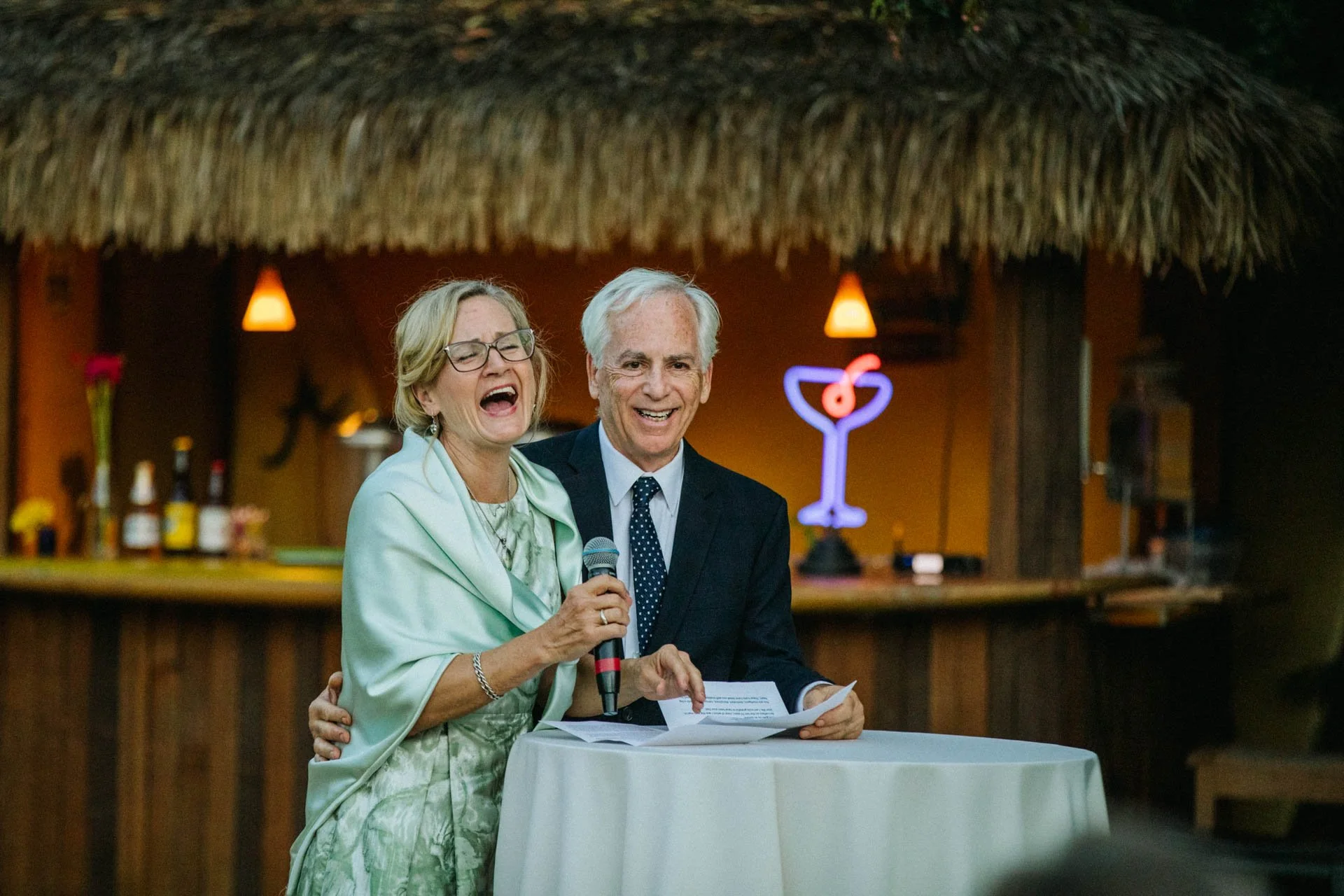 An elderly woman and man laughing and smiling at a social event, with the woman holding a microphone, standing together at a round table with a white cloth, in a tropical-themed bar or restaurant with a thatched roof and neon cocktail sign in the bac