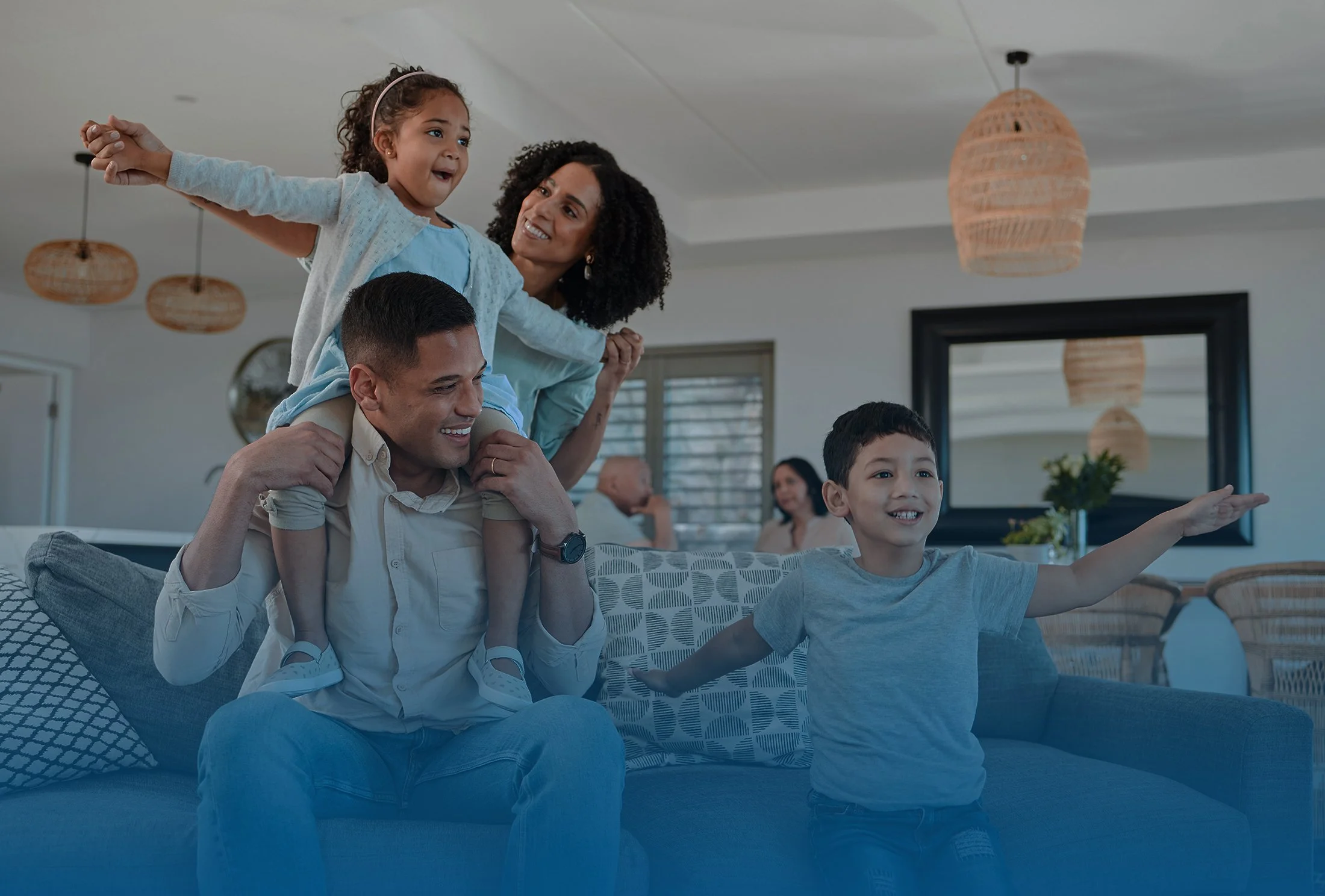A family of four enjoying time together in their living room, with a girl sitting on her father's shoulders, pointing at something out of frame, and the mother looking on smiling, while a boy sitting on the couch points as well.