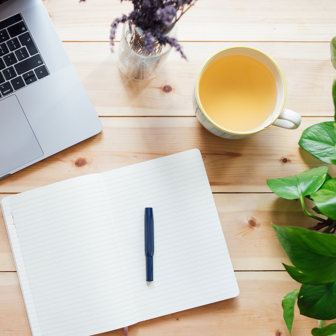 An overhead flat lay view of a peaceful wooden desk featuring an open lined notebook with a pen, a warm cup of herbal tea, a laptop, and a small vase of dried lavender.