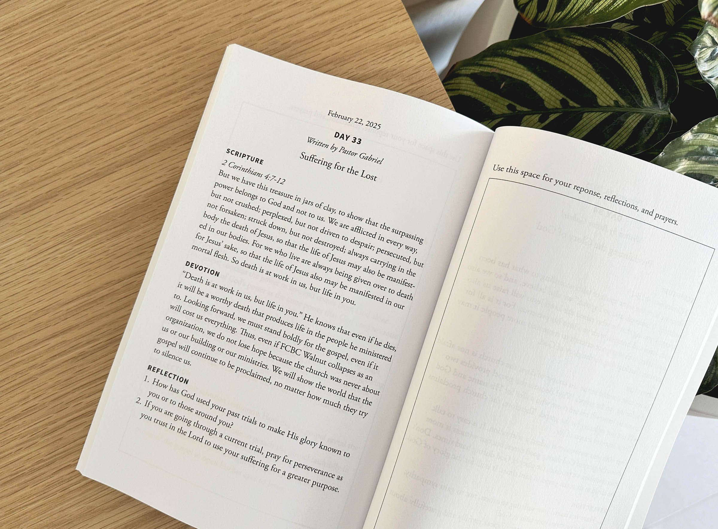 Open book on a wooden surface with a plant in the background. The page shows a date, a title, scripture reference, and devotional text.
