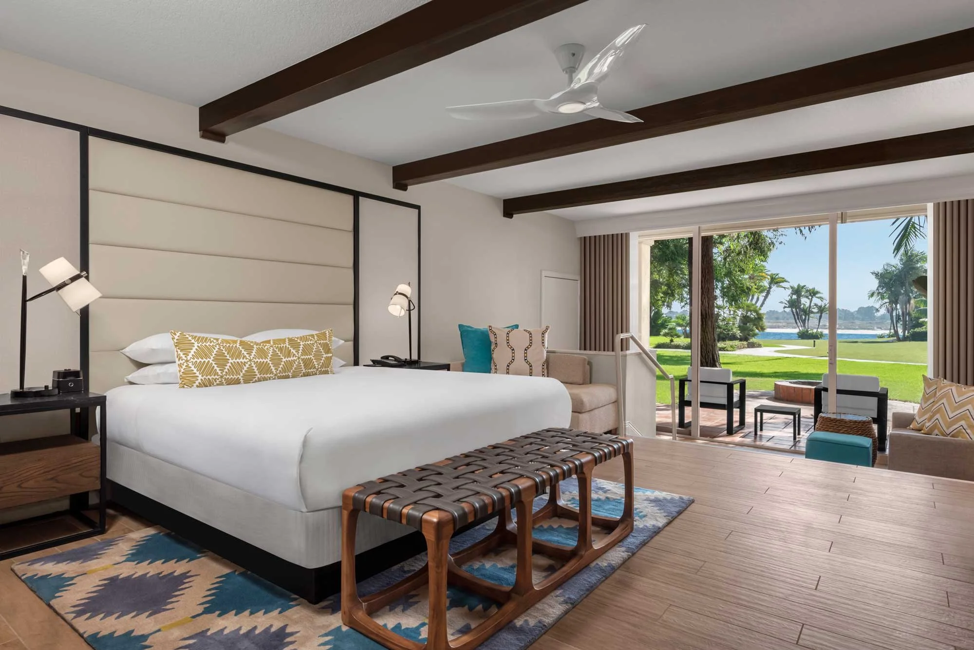 Hotel room with a large white bed, decorative pillows, bedside tables with lamps, a woven bench, seating area with chairs and a view of a green outdoor area with trees and water through sliding glass doors.