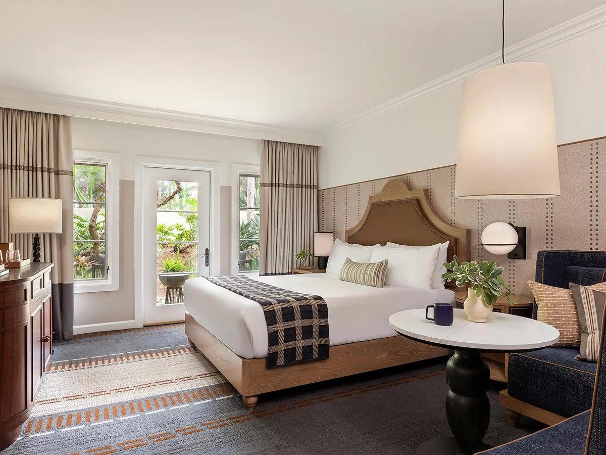 A hotel room with a king-sized bed, nightstands, lamps, a small round table with a plant and mug, a navy armchair, and large windows with beige curtains showing greenery outside.