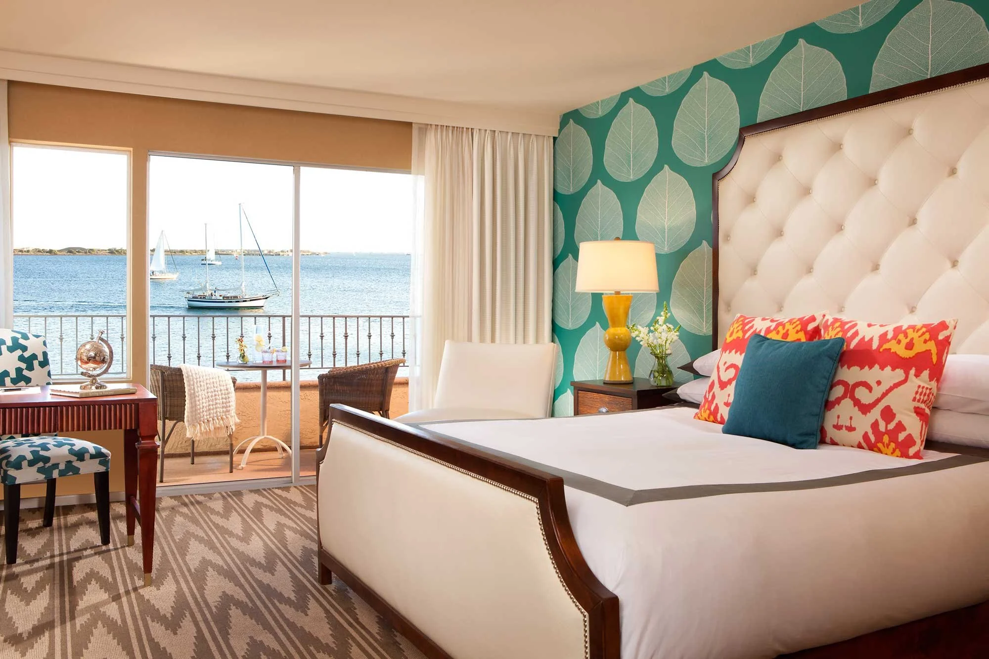 Hotel room with a view of the ocean, featuring a bed with colorful pillows, a nightstand with a lamp and flowers, a white chair, and a sliding door leading to a balcony with sailboats on the water.