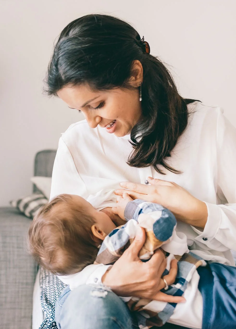 A woman breastfeeding a baby while sitting on a gray armchair in a living room.