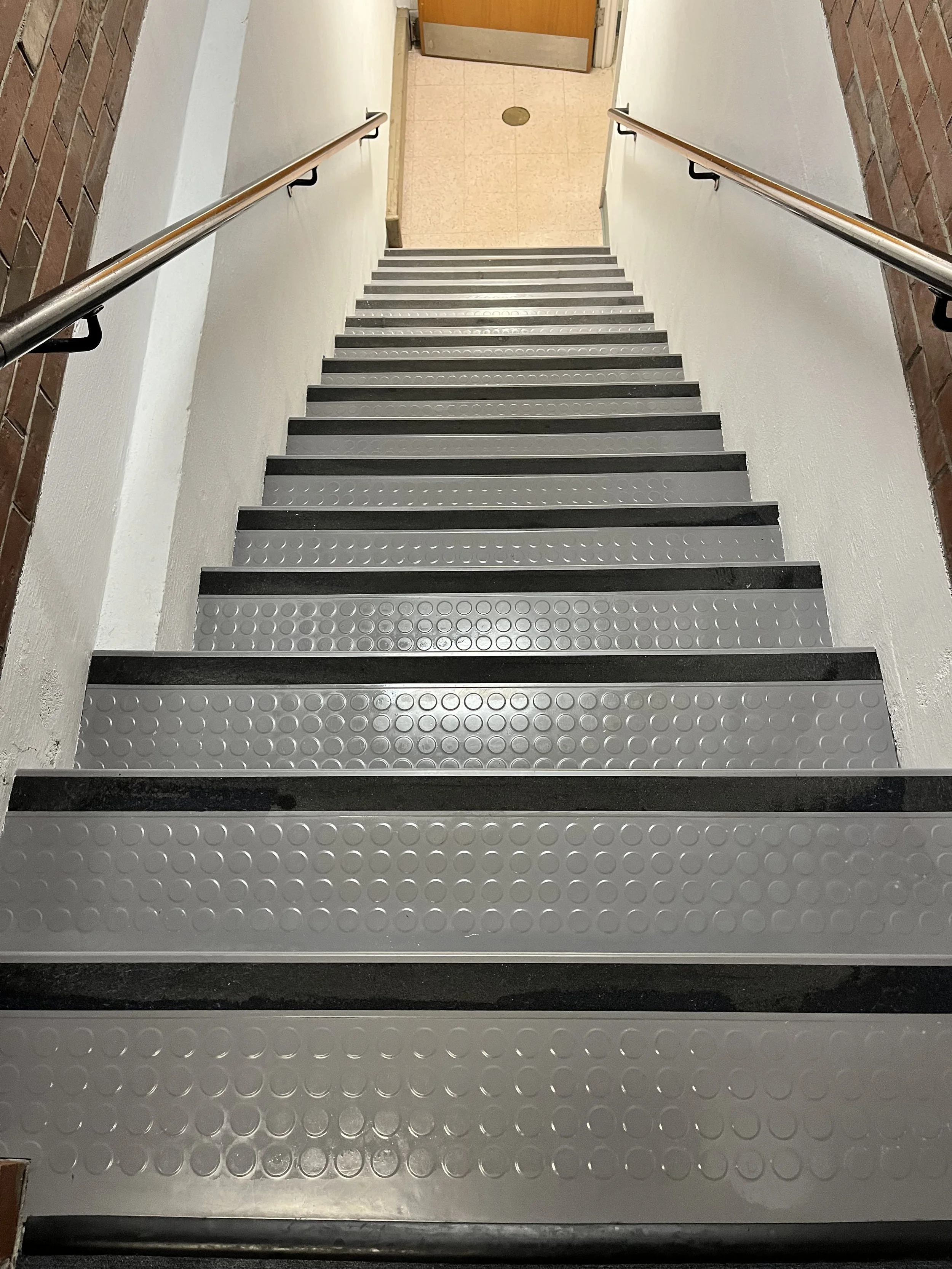 Top-down view of metal stairs with black edges and round textured surface, bordered by white walls, and supported by wooden and brick walls on either side, leading to a door at the top.