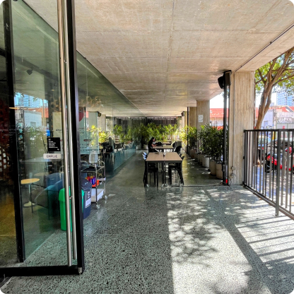 Terraza de un café moderno con mesas y sillas, plantas alrededor y un gran ventanal. Personas sentadas en las mesas dentro del establecimiento.