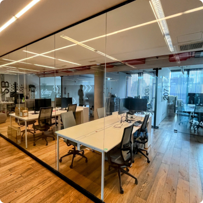 Oficina moderna con paredes de vidrio, piso de madera, escritorios blancos y sillas ergonómicas. Hay varias computadoras en los escritorios y el espacio está bien iluminado con luz natural y fluorescente.